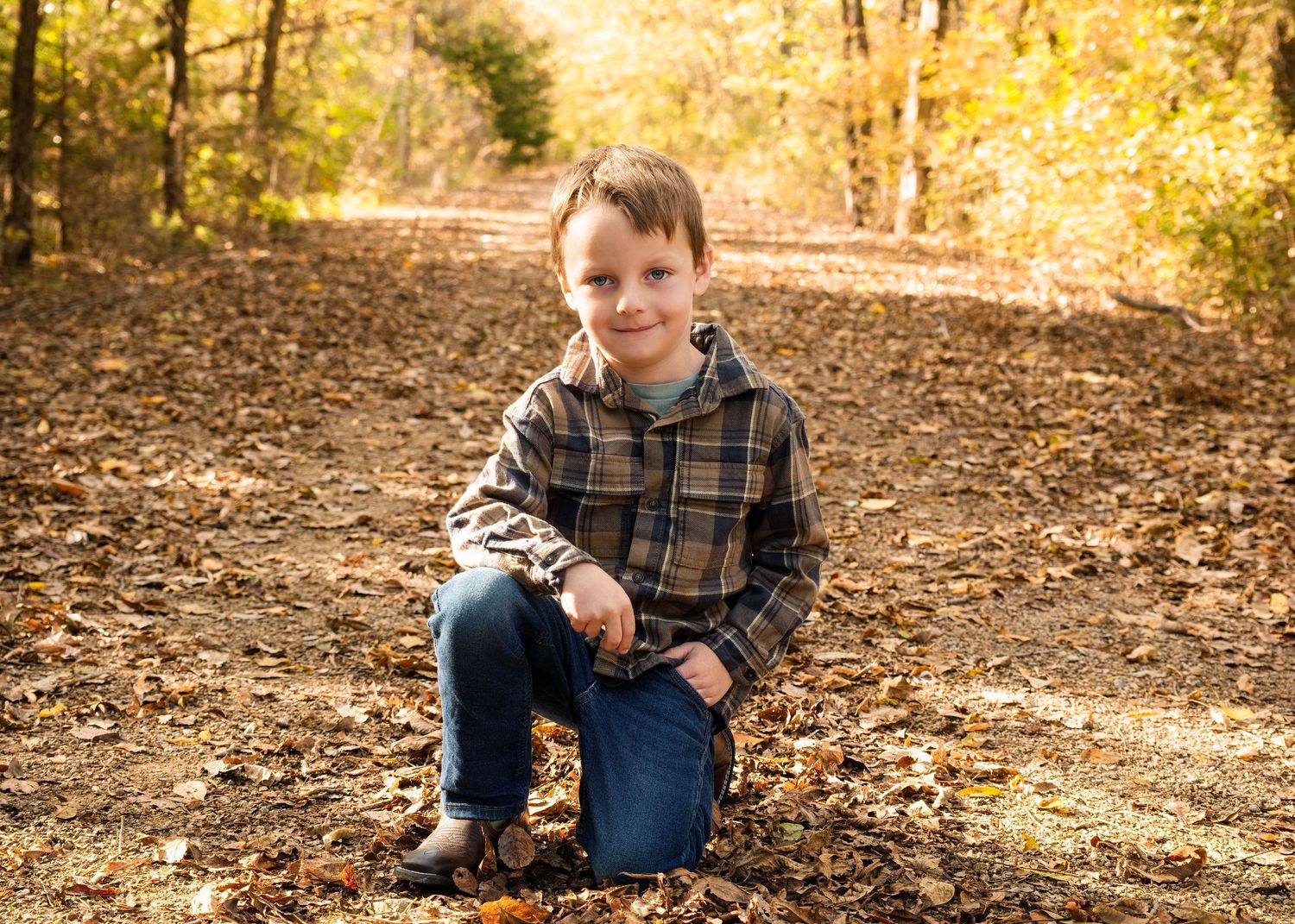 Young child kneels on fallen leaves during autumn photo session on wooded trail.