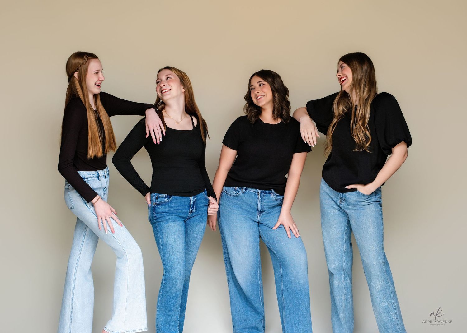 Four women in black tops and blue jeans laughing together against a neutral background.