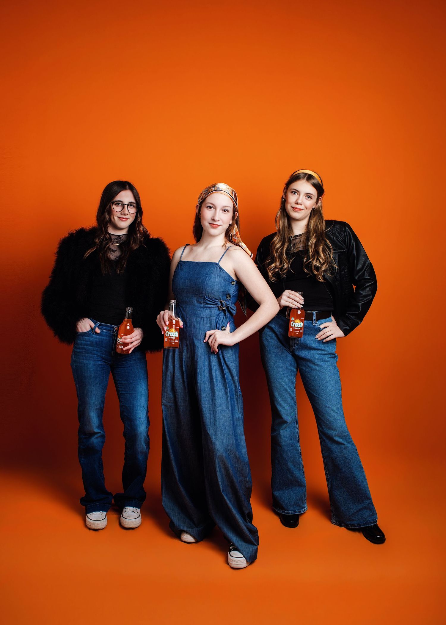 Three people in denim and dark clothing sit casually against an orange backdrop in a creative studio photoshoot.