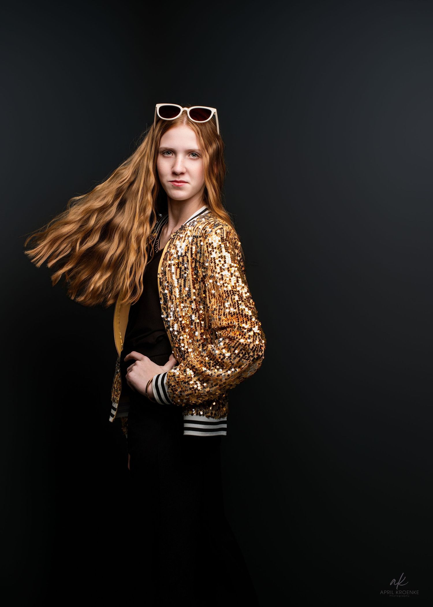 A young redhead woman poses in a gold sequin jacket with white sunglasses, her long hair flowing against a dark background.