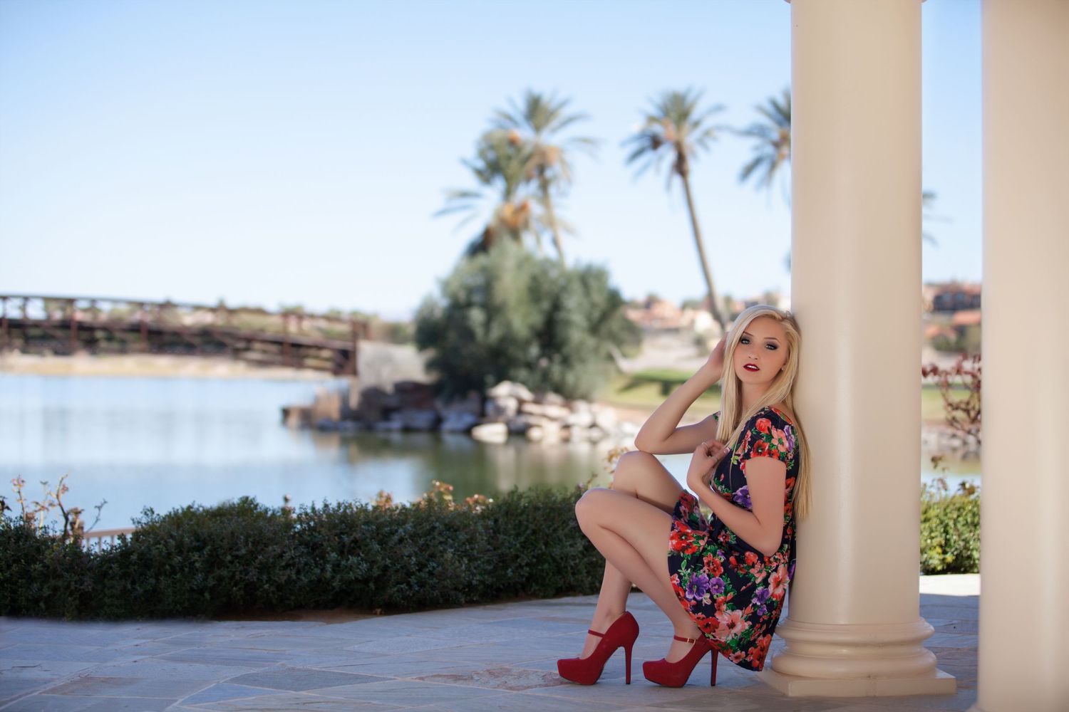 Woman in red high heels and summer dress poses elegantly by white columns overlooking a tropical waterfront.