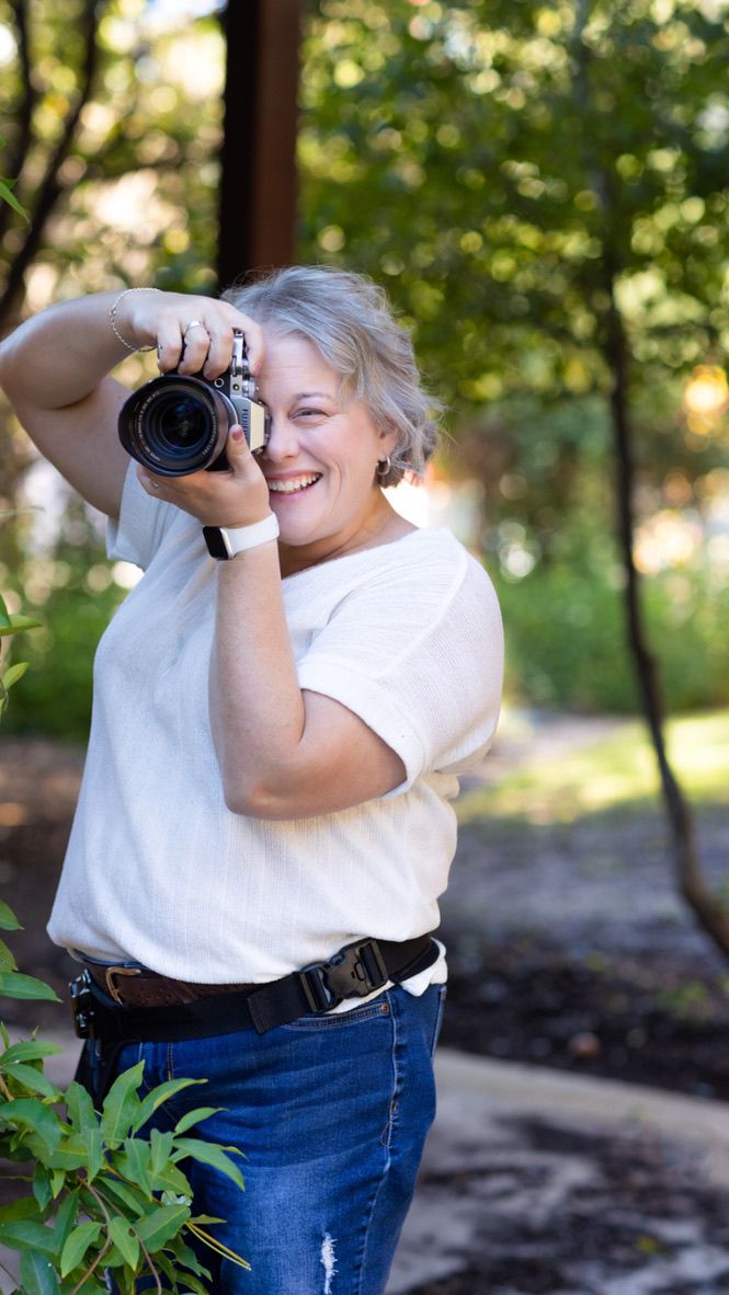 A photographer in jeans and white shirt takes photos in a sunny park setting with trees in the background.