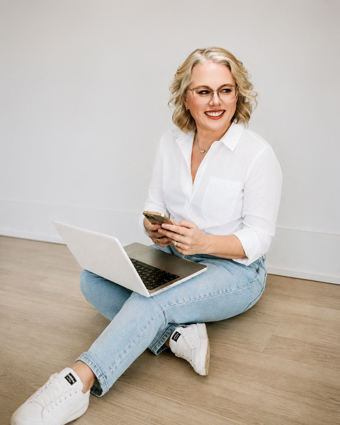 Smiling blonde woman with glasses sits on floor with laptop and phone, wearing white shirt and jeans.