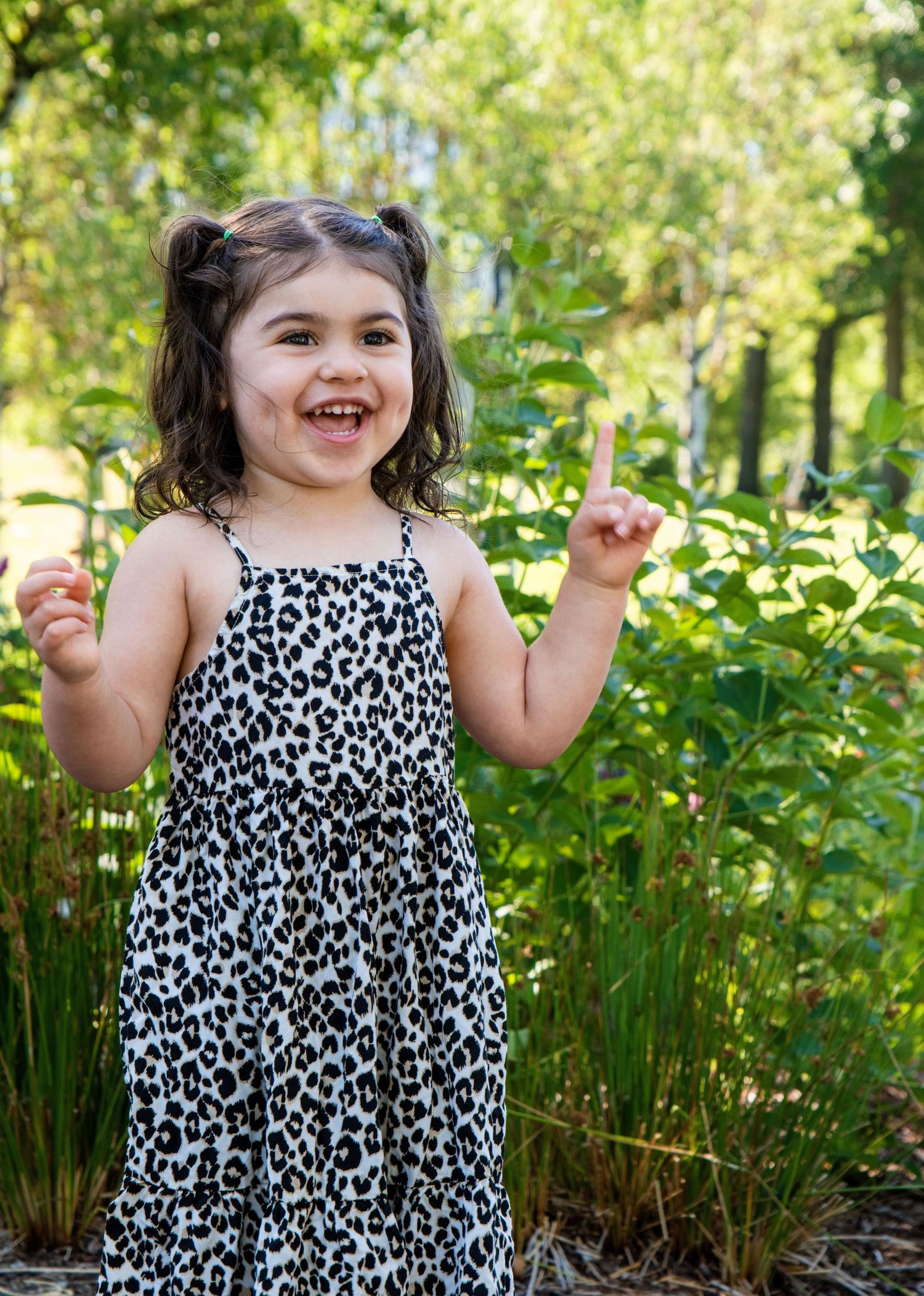 Child in leopard print dress poses cheerfully against green garden background.