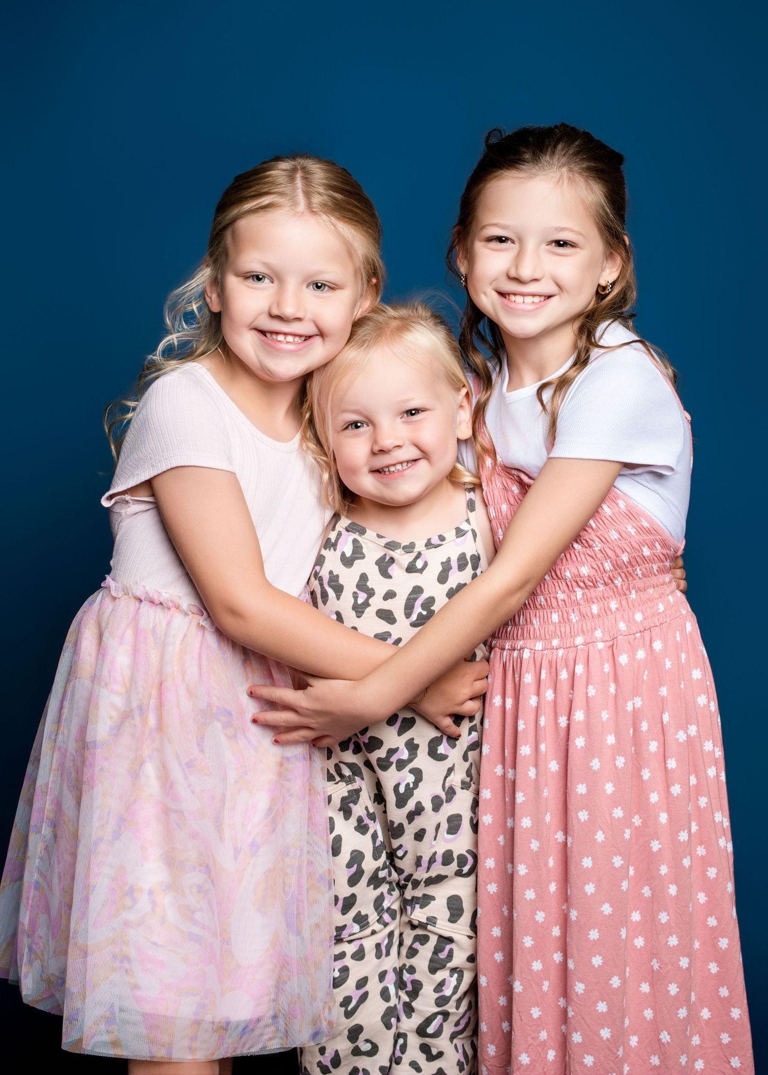 Young children in dresses hugging and smiling against blue studio background.