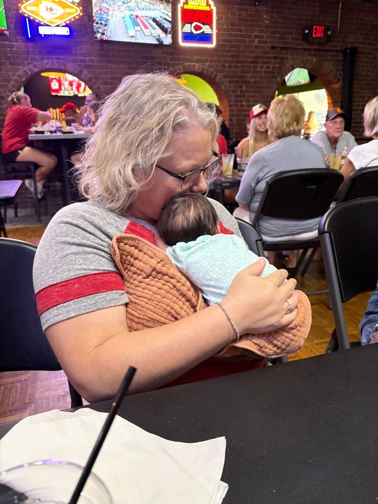 A person in a gray shirt holds a sleeping baby at a casual indoor gathering with neon signs on brick walls.