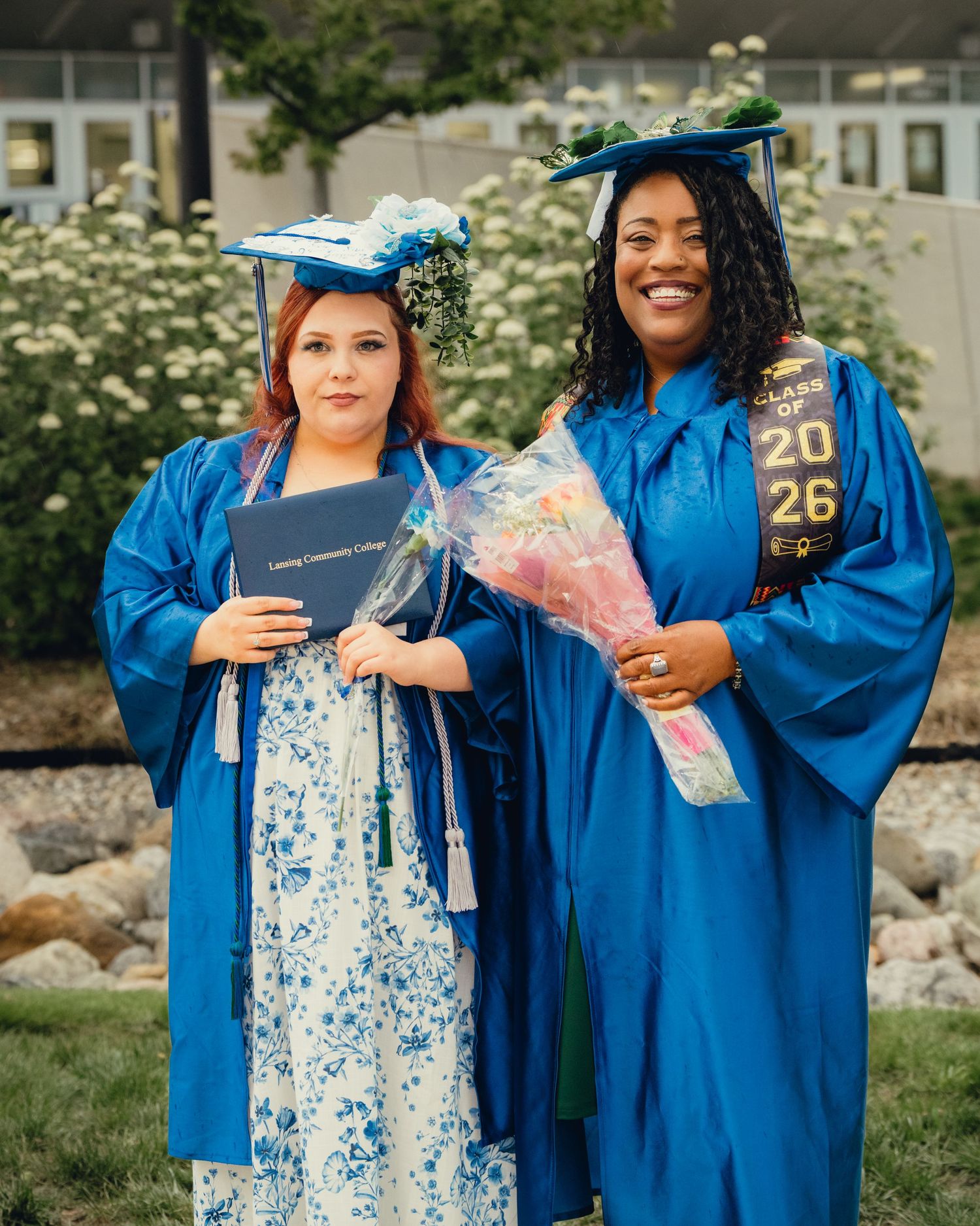 Two women in blue graduation gowns and caps smile proudly, one holding a diploma and the other a bouquet of flowers.