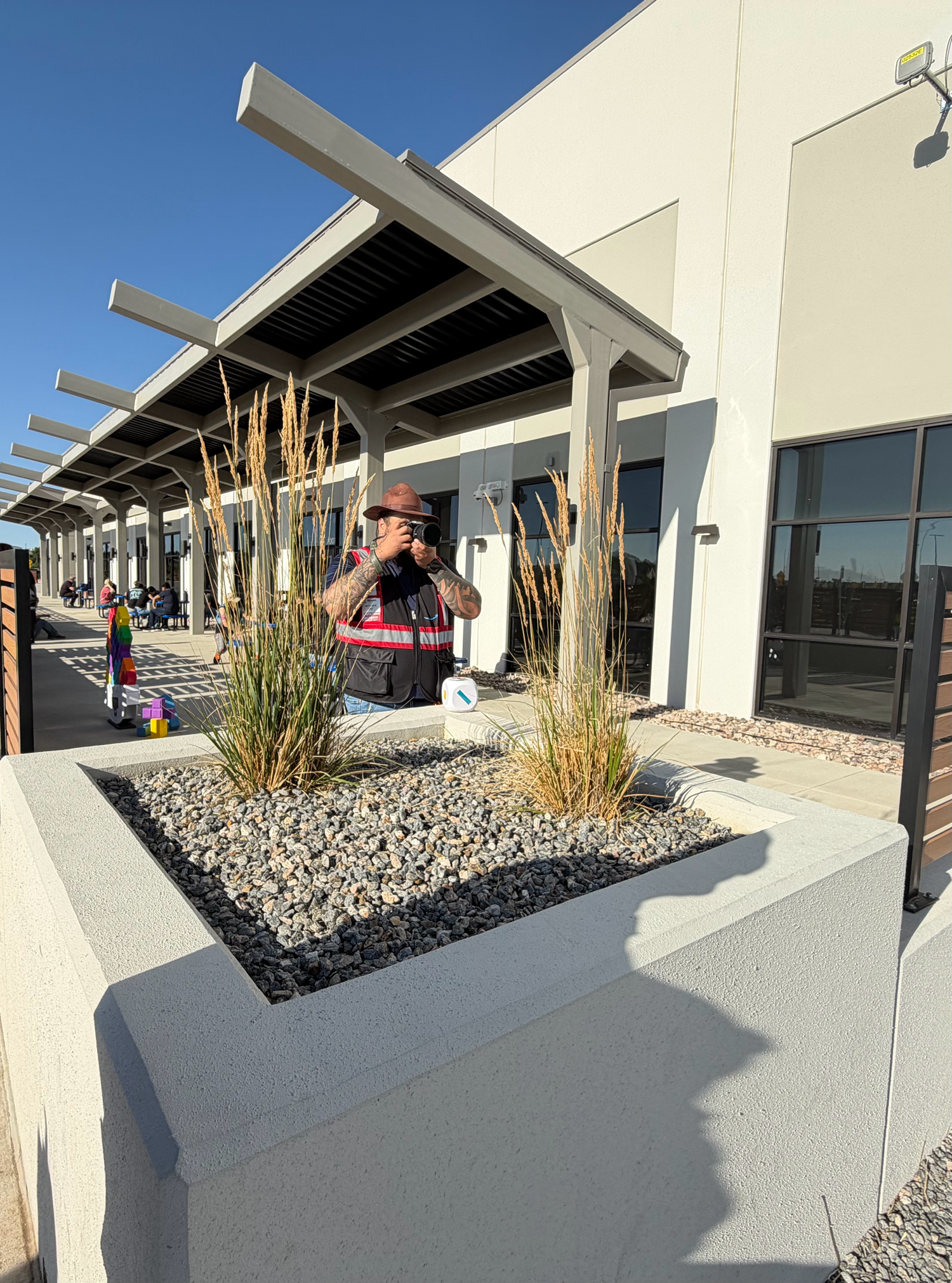 A worker in a safety vest photographs a modern building entrance with decorative gravel planters and ornamental grasses under a pergola.