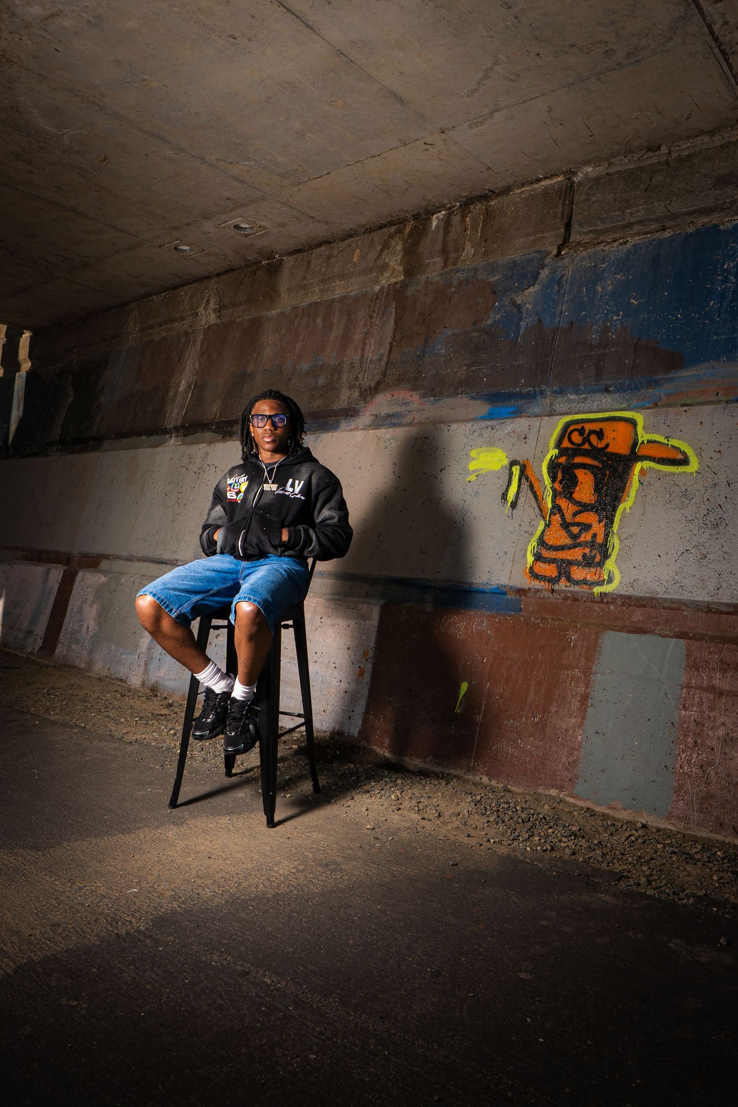Young man sitting on stool under urban bridge with colorful graffiti art on concrete wall behind him.