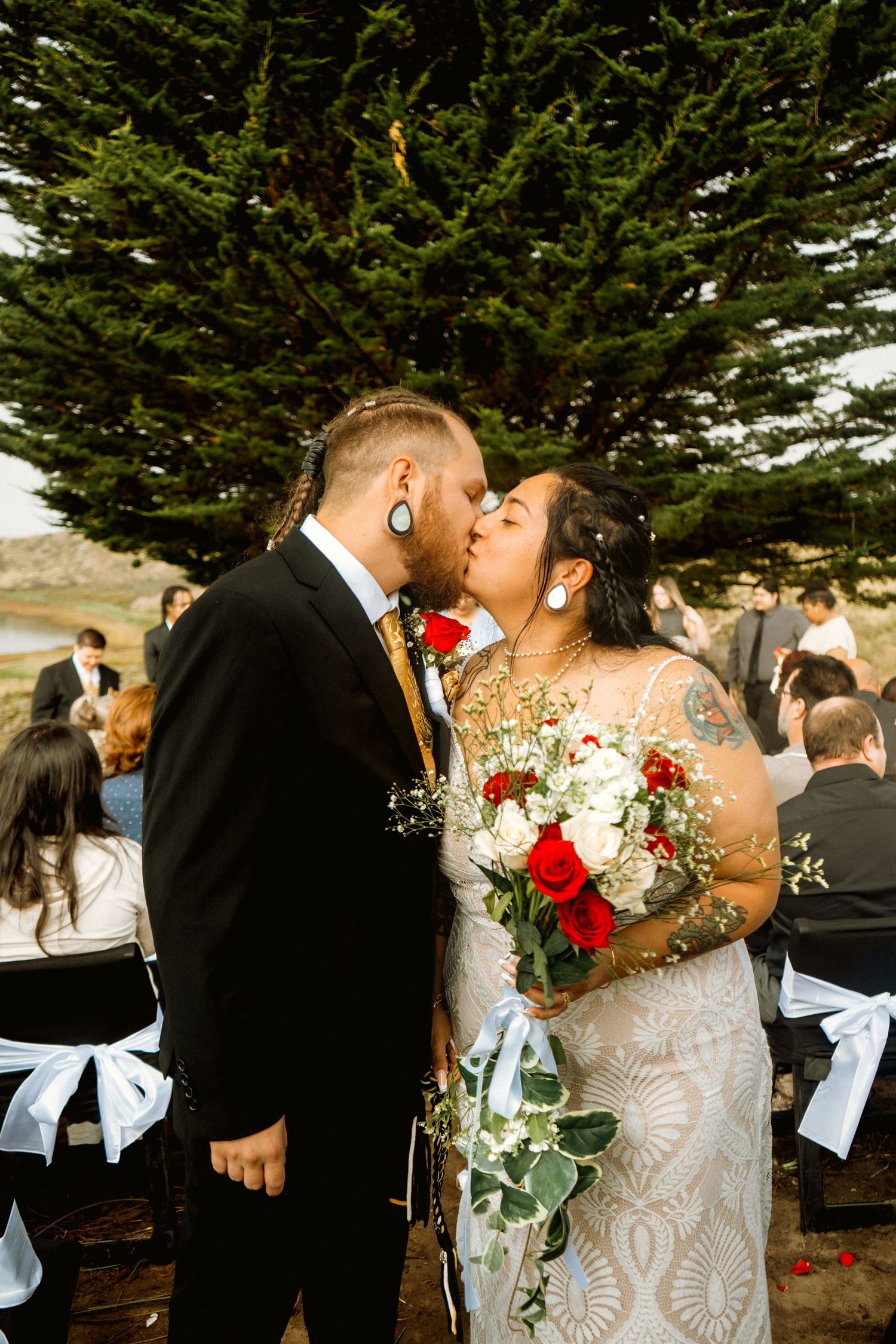 Tattooed bride and groom share first kiss holding red and white bouquet at outdoor wedding.