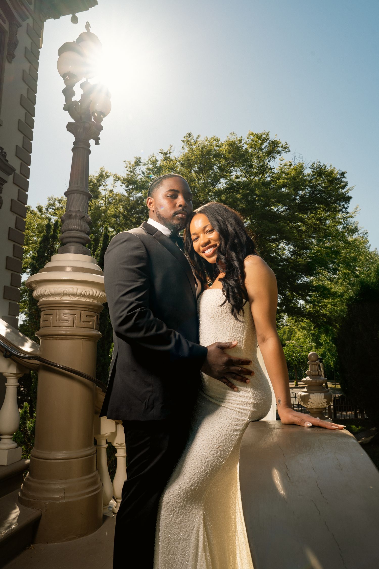 Elegant couple in wedding attire pose near ornate lamppost with sunlight behind them.