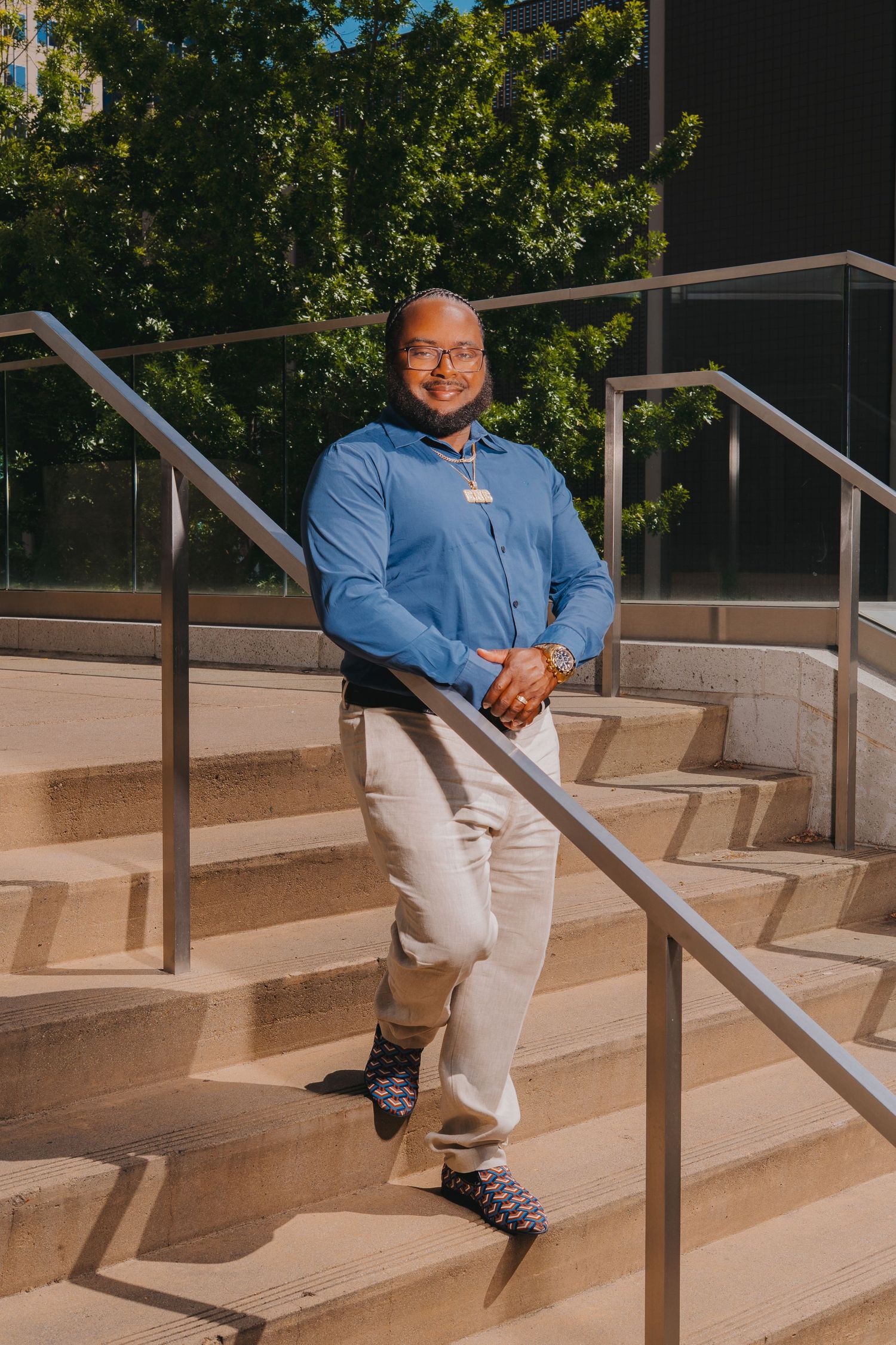 Young man in blue shirt and khaki pants poses on outdoor steps near modern glass building.