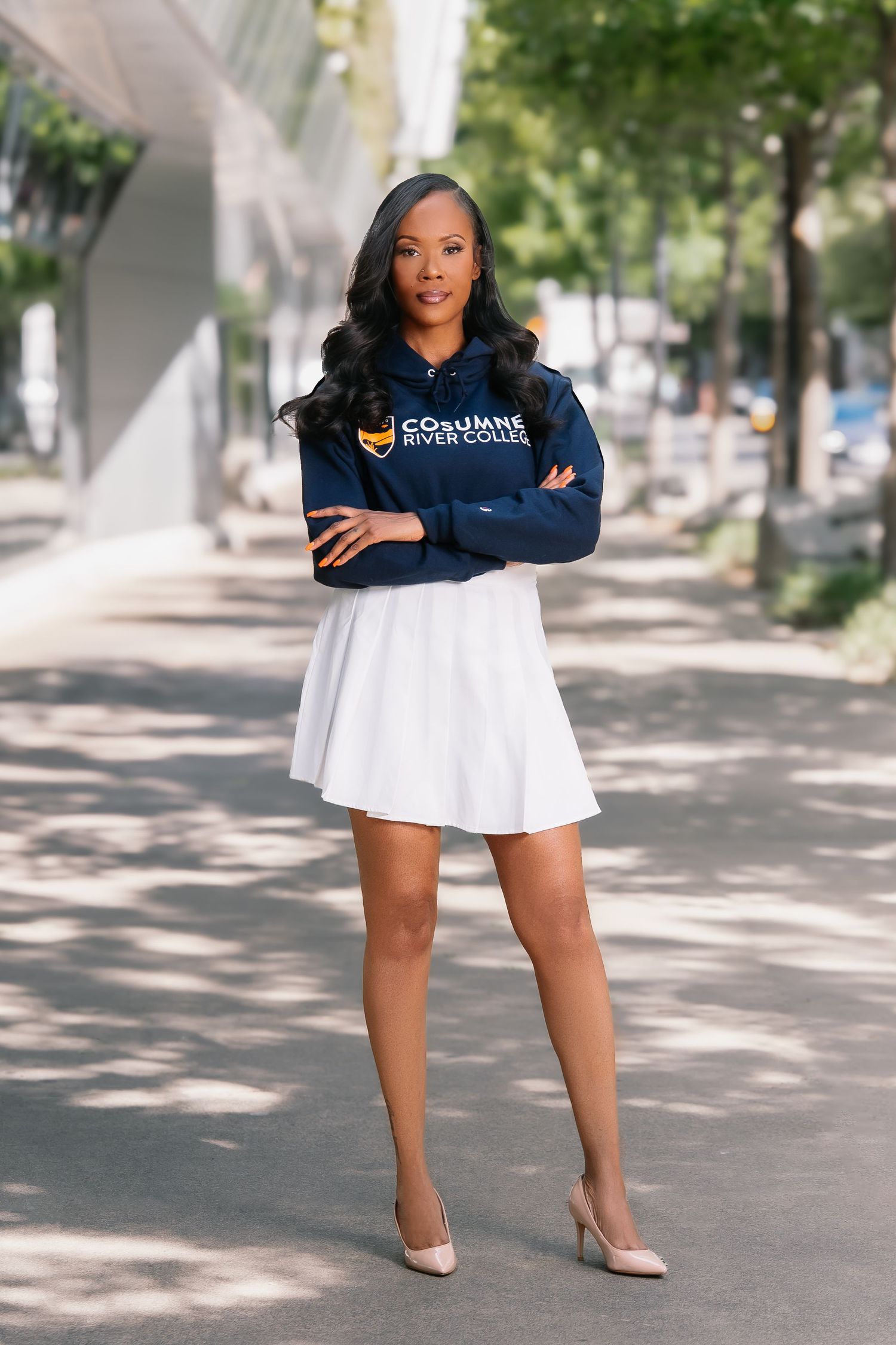 Young Black woman in Cosumnes River College hoodie and white skirt posing outdoors with arms crossed.