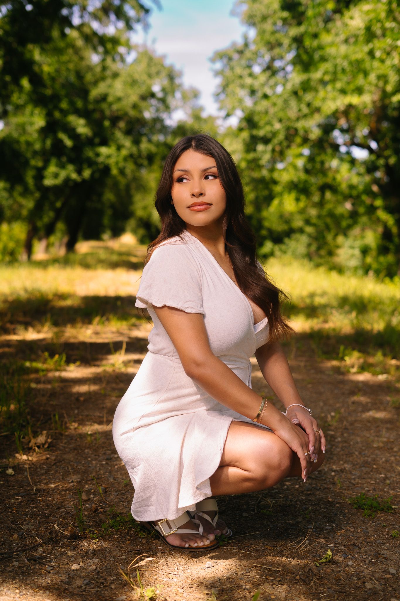 A young woman in a white dress crouches gracefully on a dirt path surrounded by lush green trees.