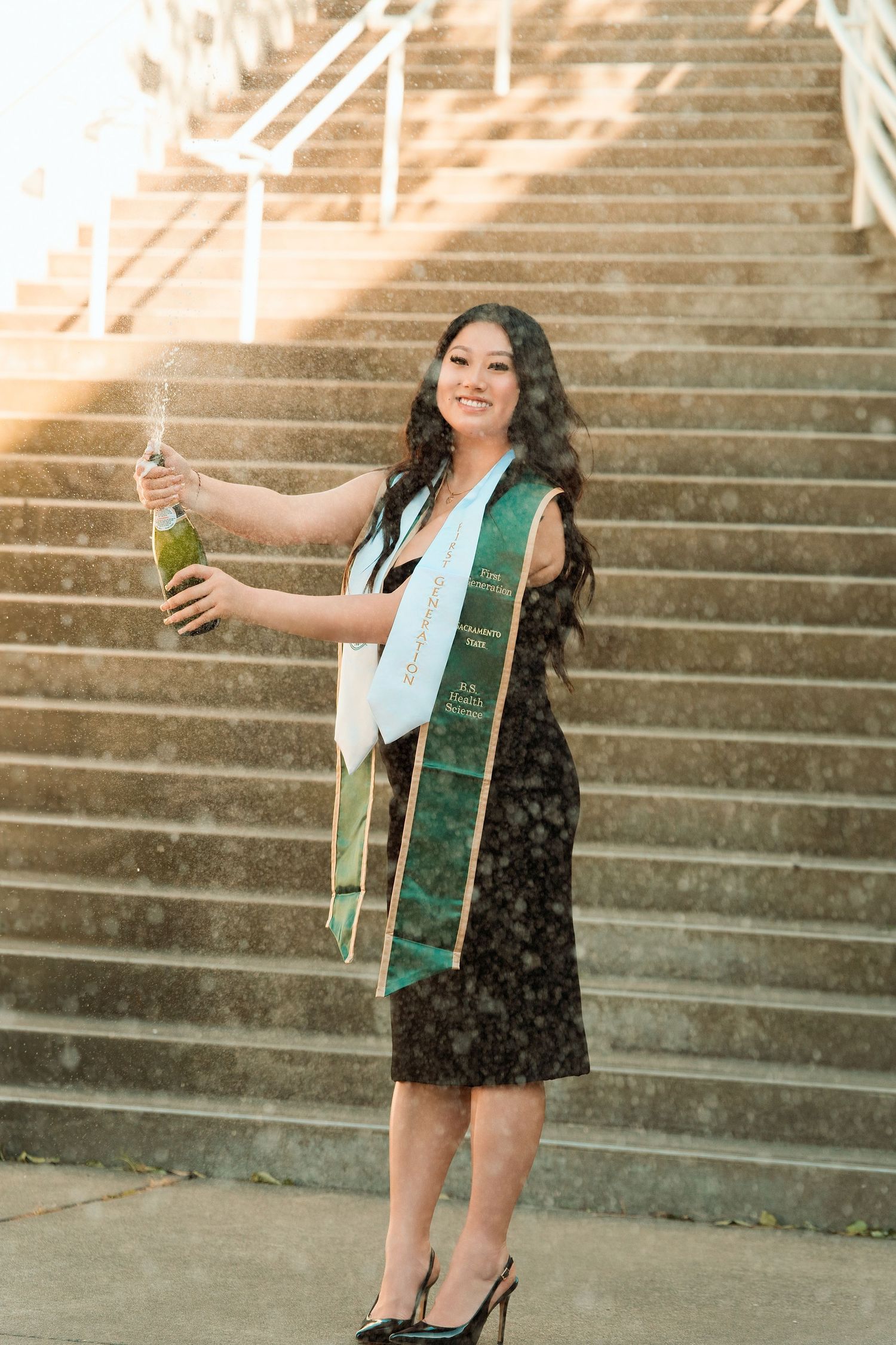 Graduate in black dress with green sash pops champagne bottle on outdoor staircase during celebration.