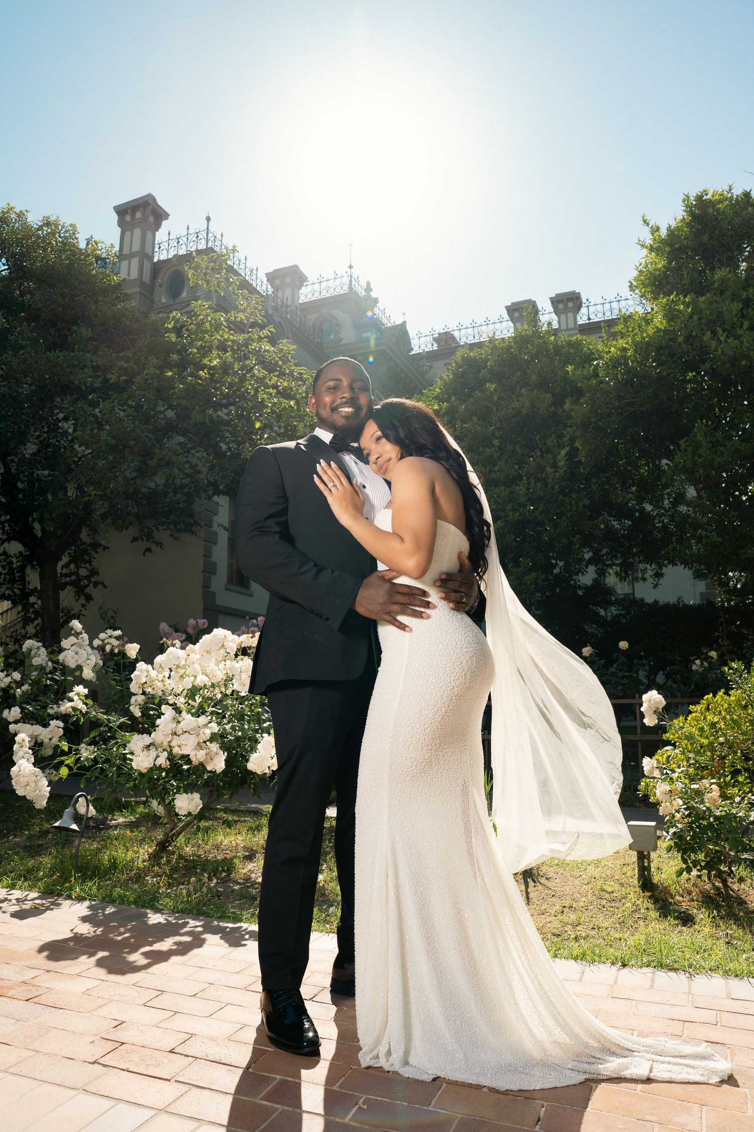 Groom in black tuxedo embraces bride in white gown near white roses in sunlight.