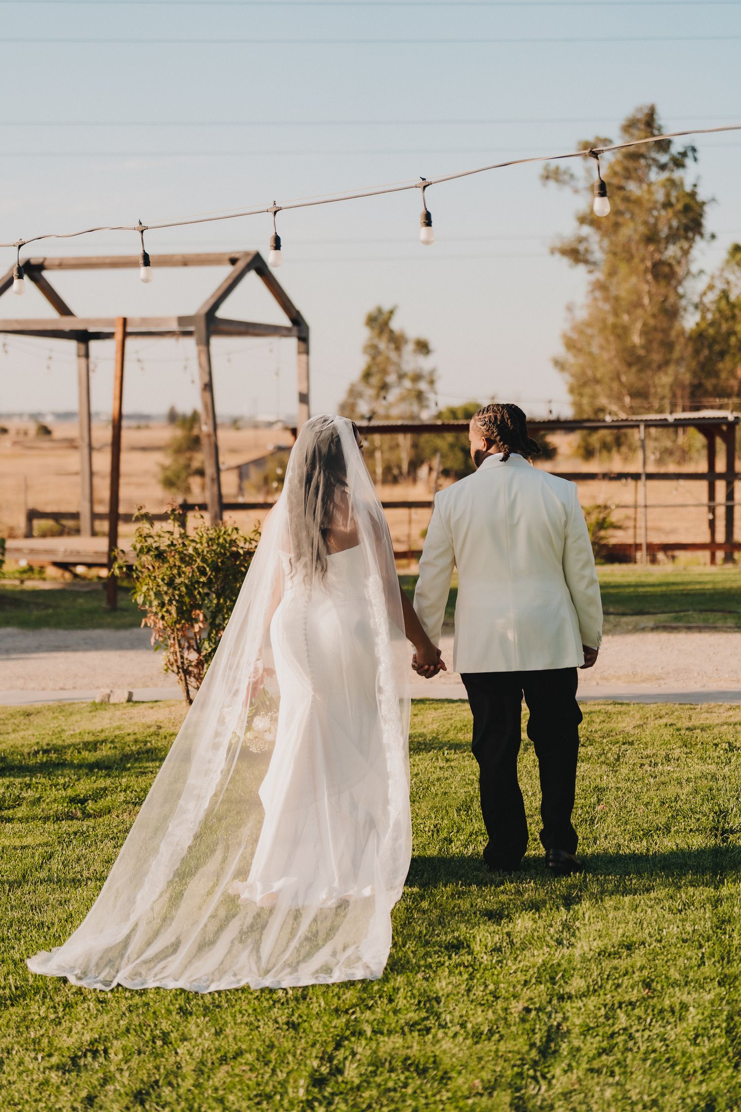 Bride and groom holding hands walking across green lawn at outdoor rustic wedding venue with string lights.