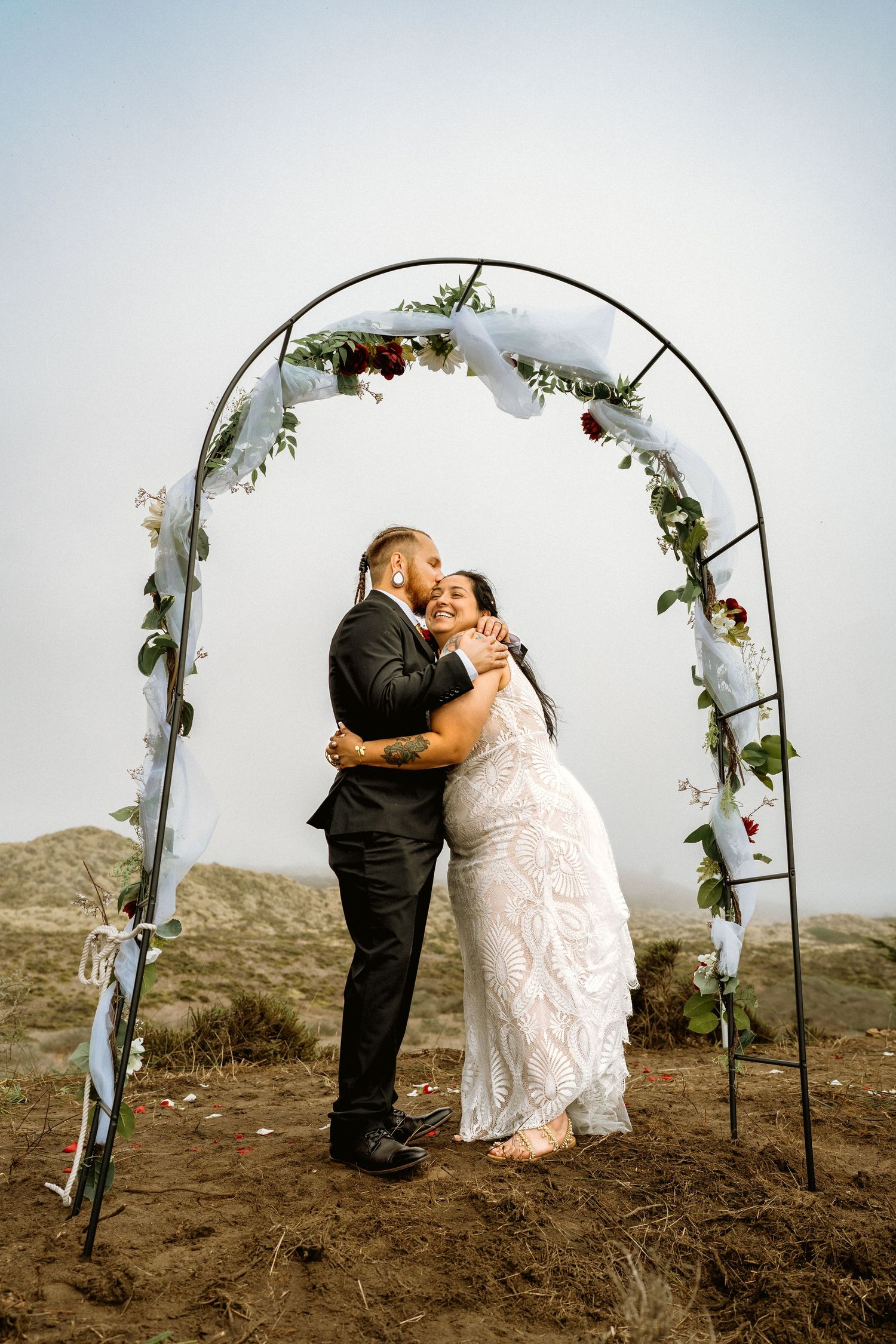 Newlywed couple shares a joyful embrace under a floral arch on a foggy coastal hillside.