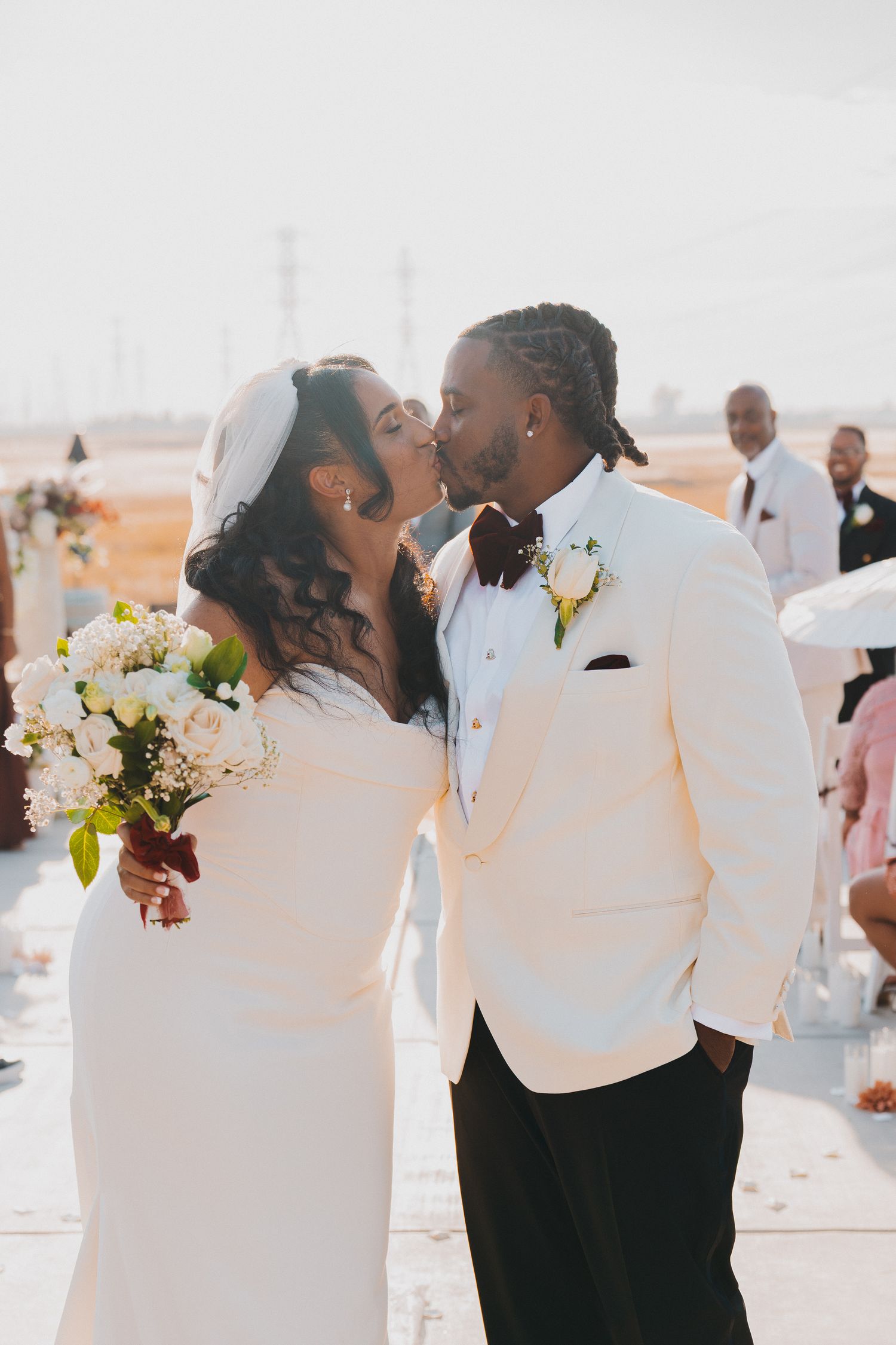 Bride and groom sharing first kiss at outdoor wedding ceremony while bride holds white floral bouquet.