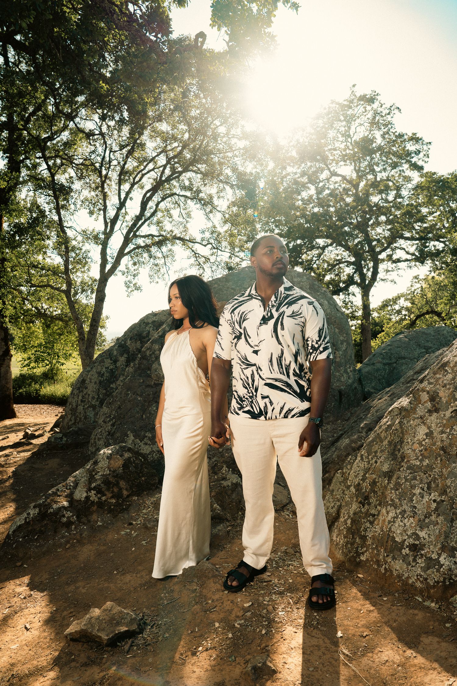 Couple in white outfits stand among large rocks under sunlit trees in nature.