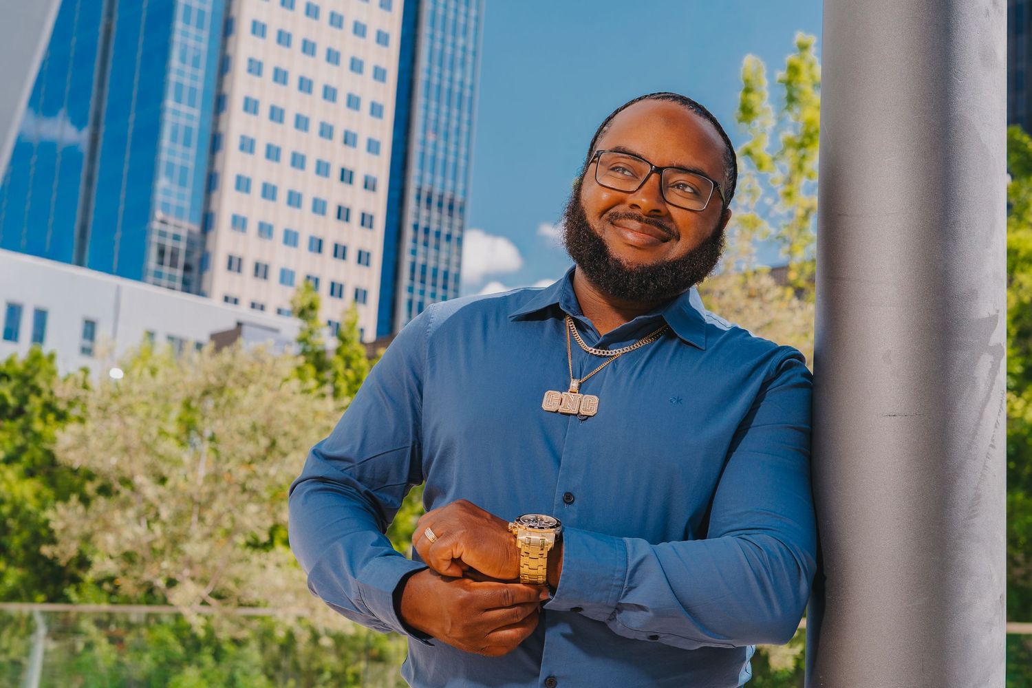 Smiling young man in blue shirt holds drink while leaning against pole with city buildings in background.