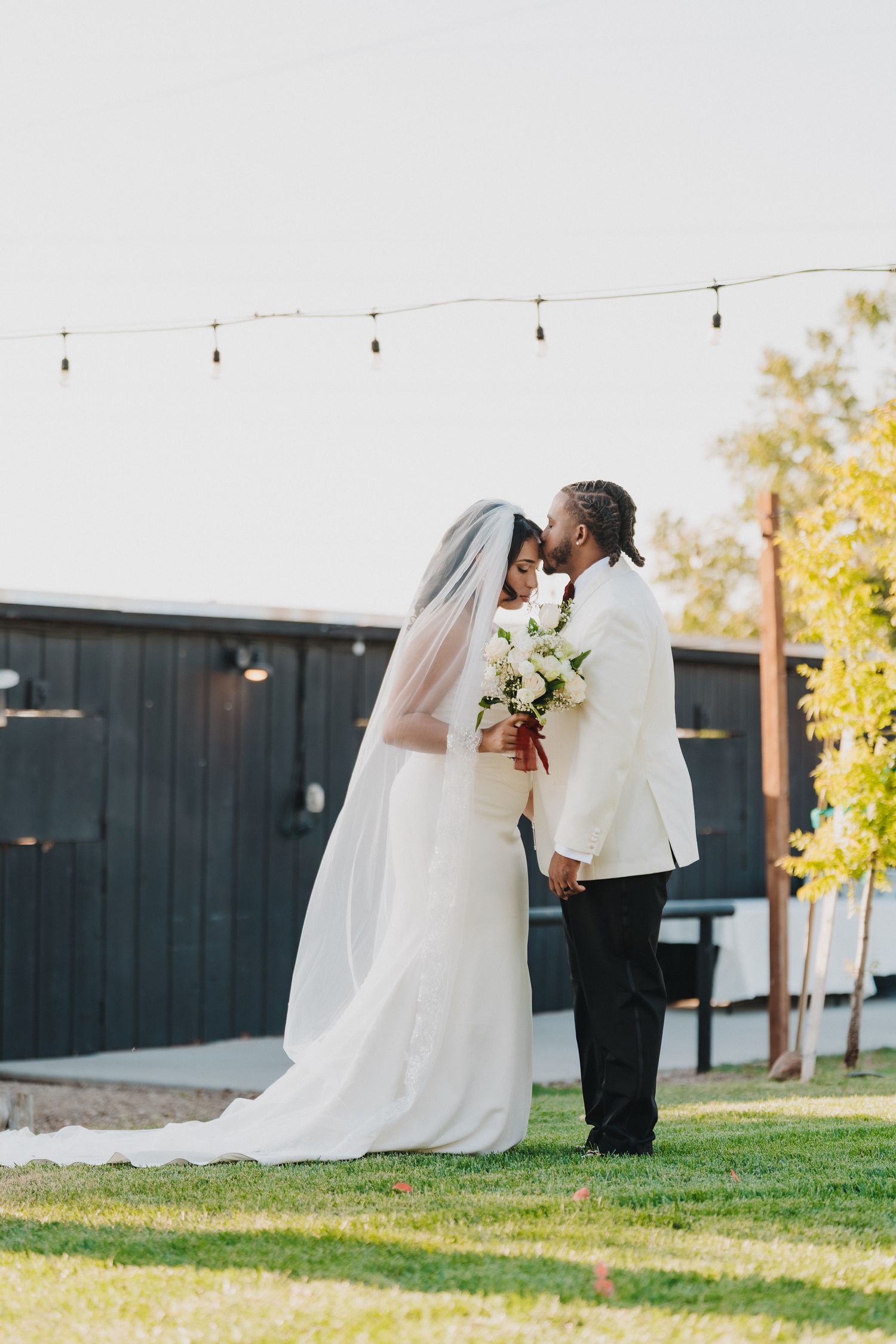 Bride and groom sharing romantic kiss on green lawn under string lights at outdoor wedding venue.