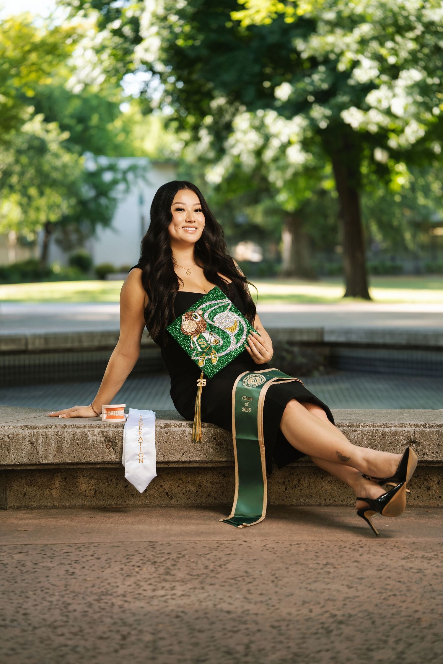 Smiling graduate in black dress holds decorated cap, wearing sash, seated outdoors on stone steps with trees in background.