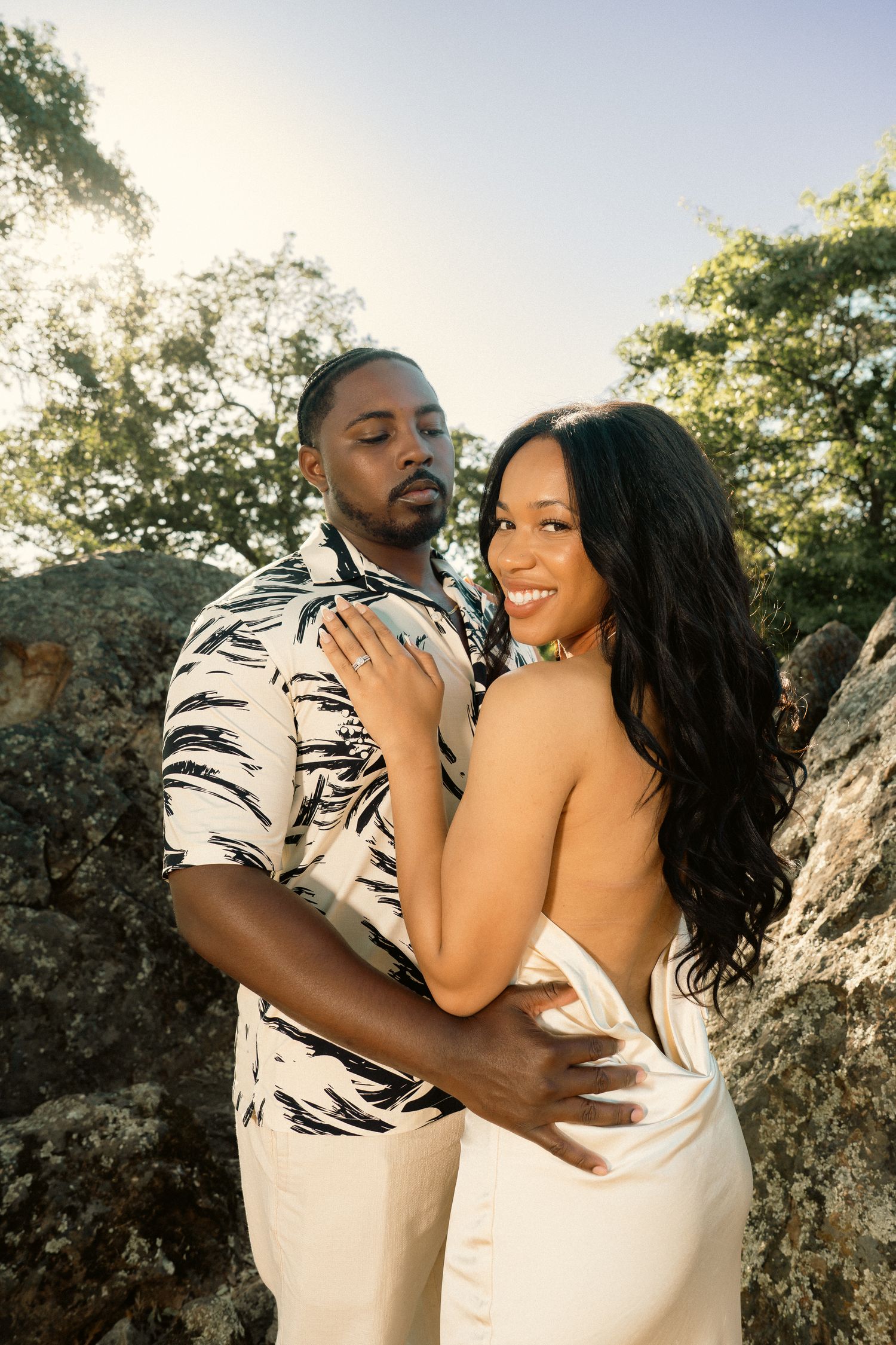 Couple embraces near rocky outcrop at sunset, woman in backless white dress smiling.
