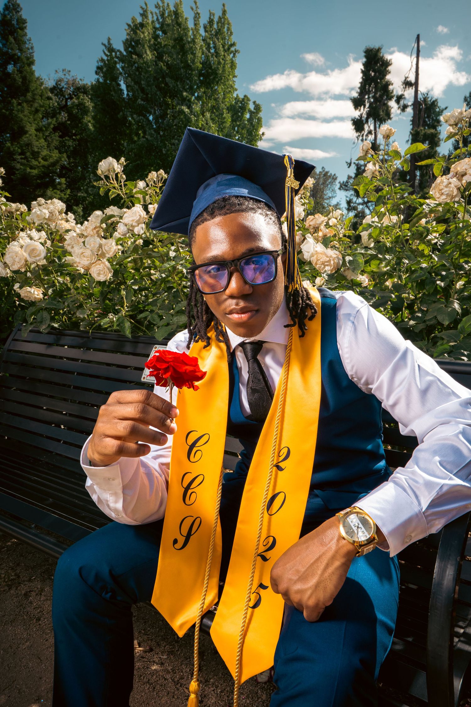 Graduate in blue vest and gold stole holding red rose while seated on bench.