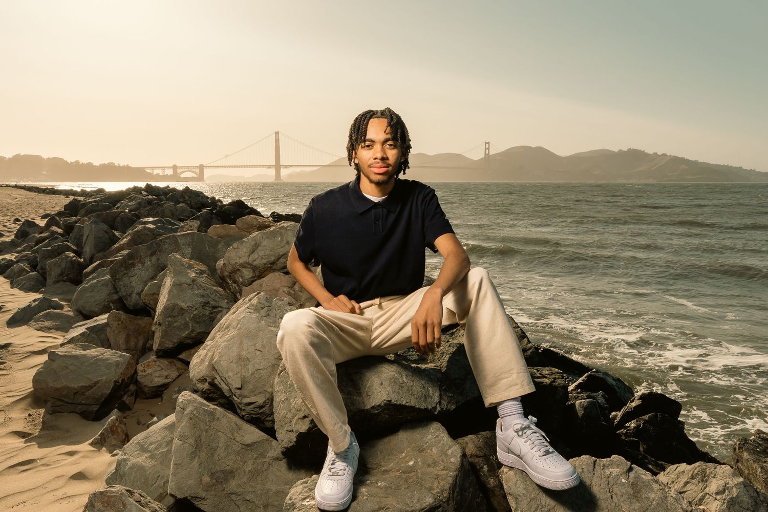 Young man in navy shirt and cream pants sitting on rocks with Golden Gate Bridge backdrop.