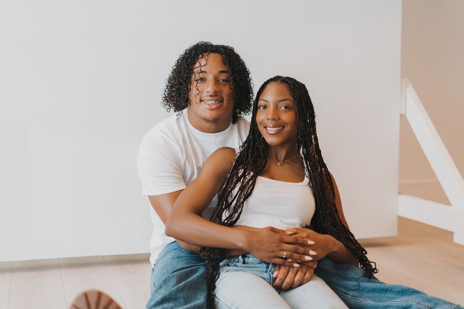 Smiling young couple sits closely together in casual white outfits in bright room.
