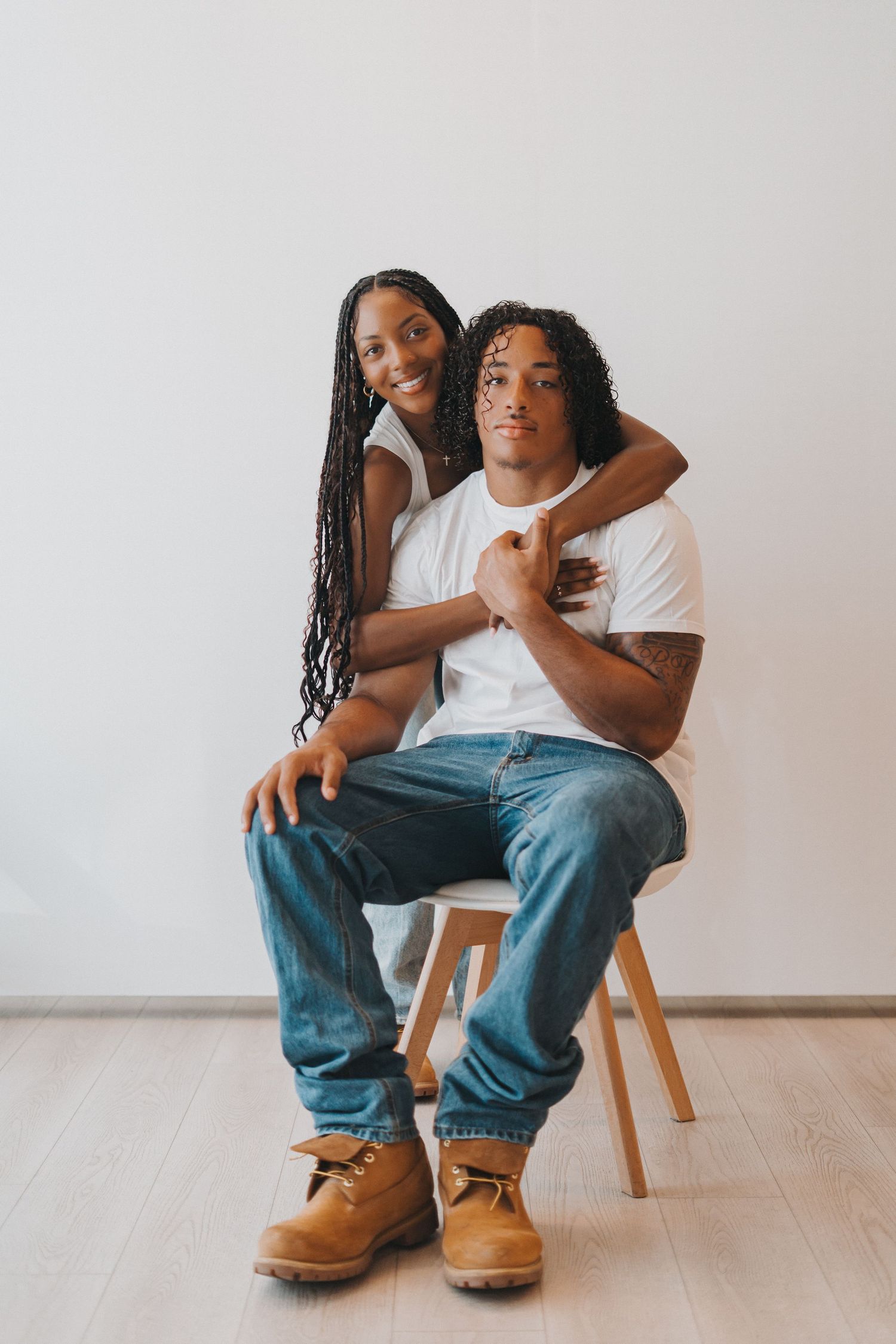 Young couple in white tees and jeans pose together in bright minimalist studio setting.