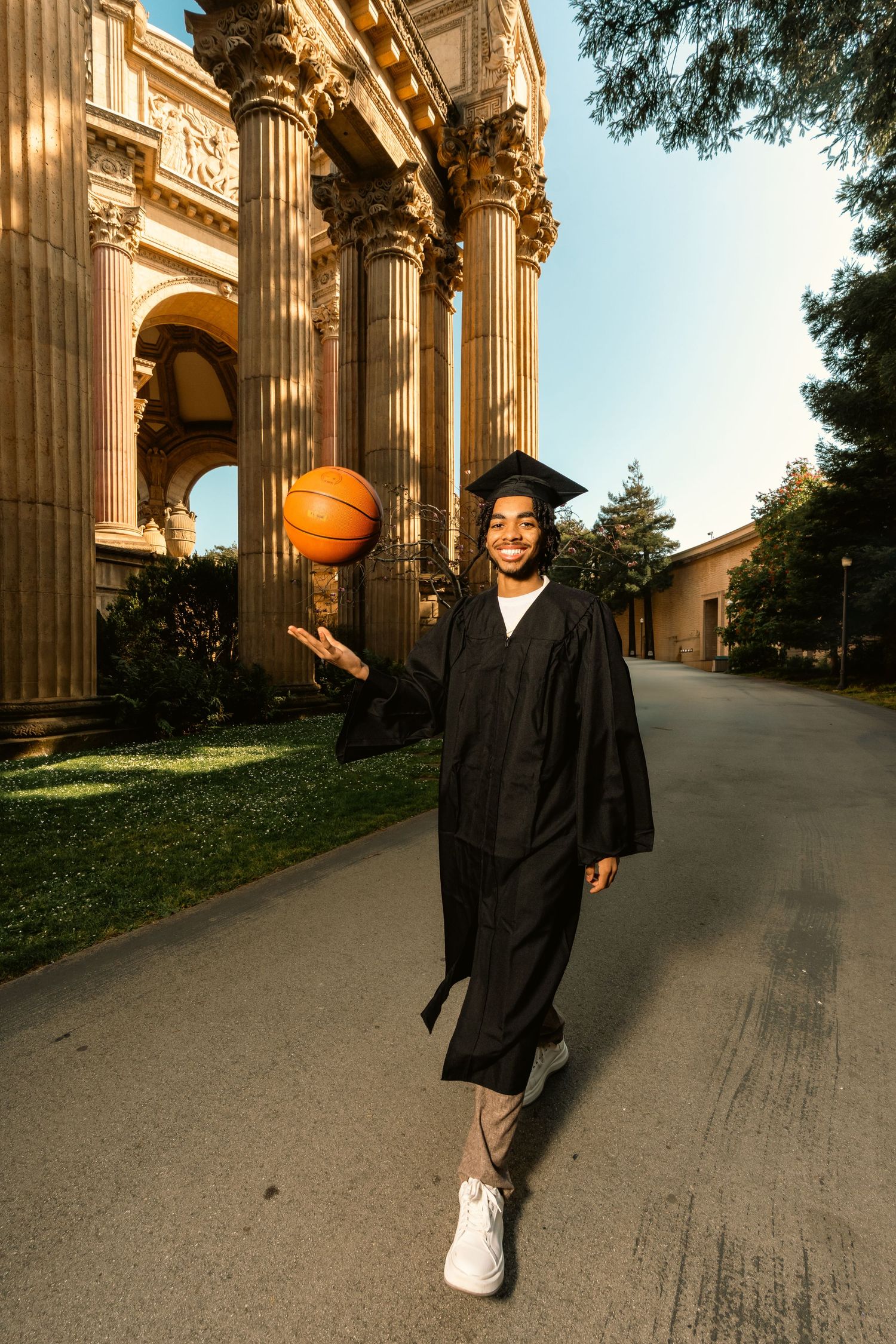 Smiling graduate in black cap and gown spinning basketball outside ornate classical building.