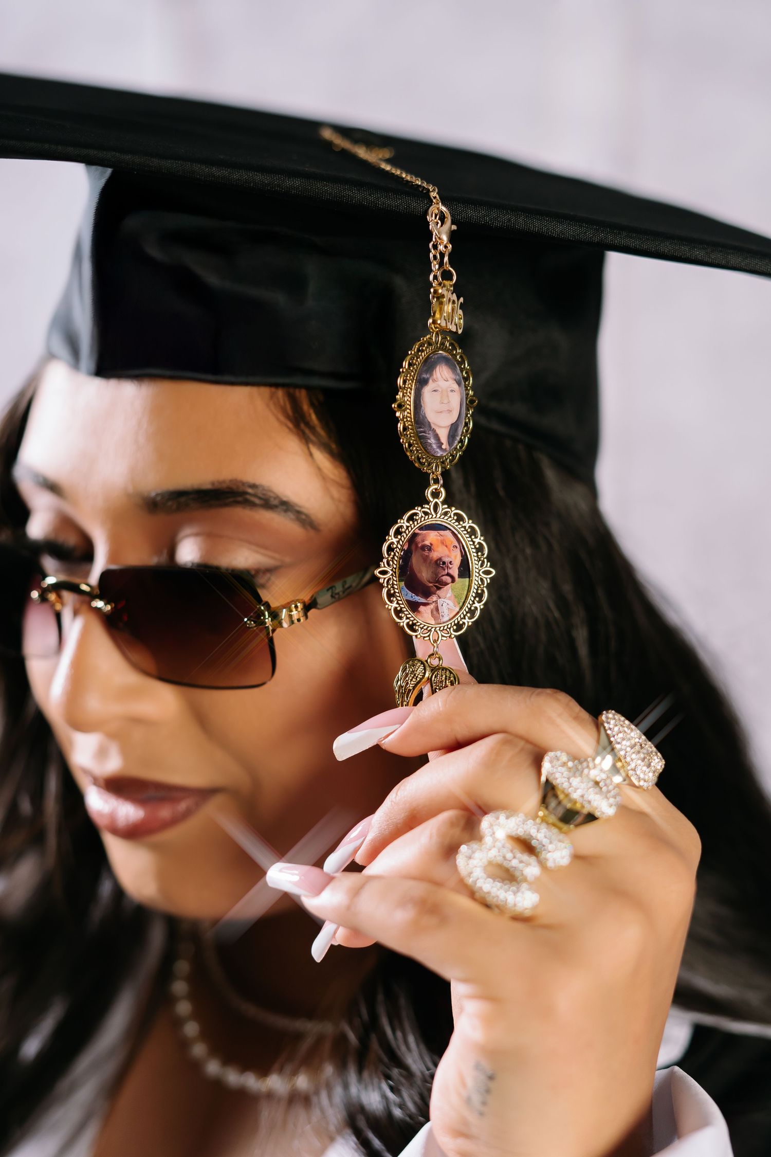 Graduate in black cap with memorial photo charms, sunglasses, pearl necklace, and rhinestone rings poses for graduation photo.