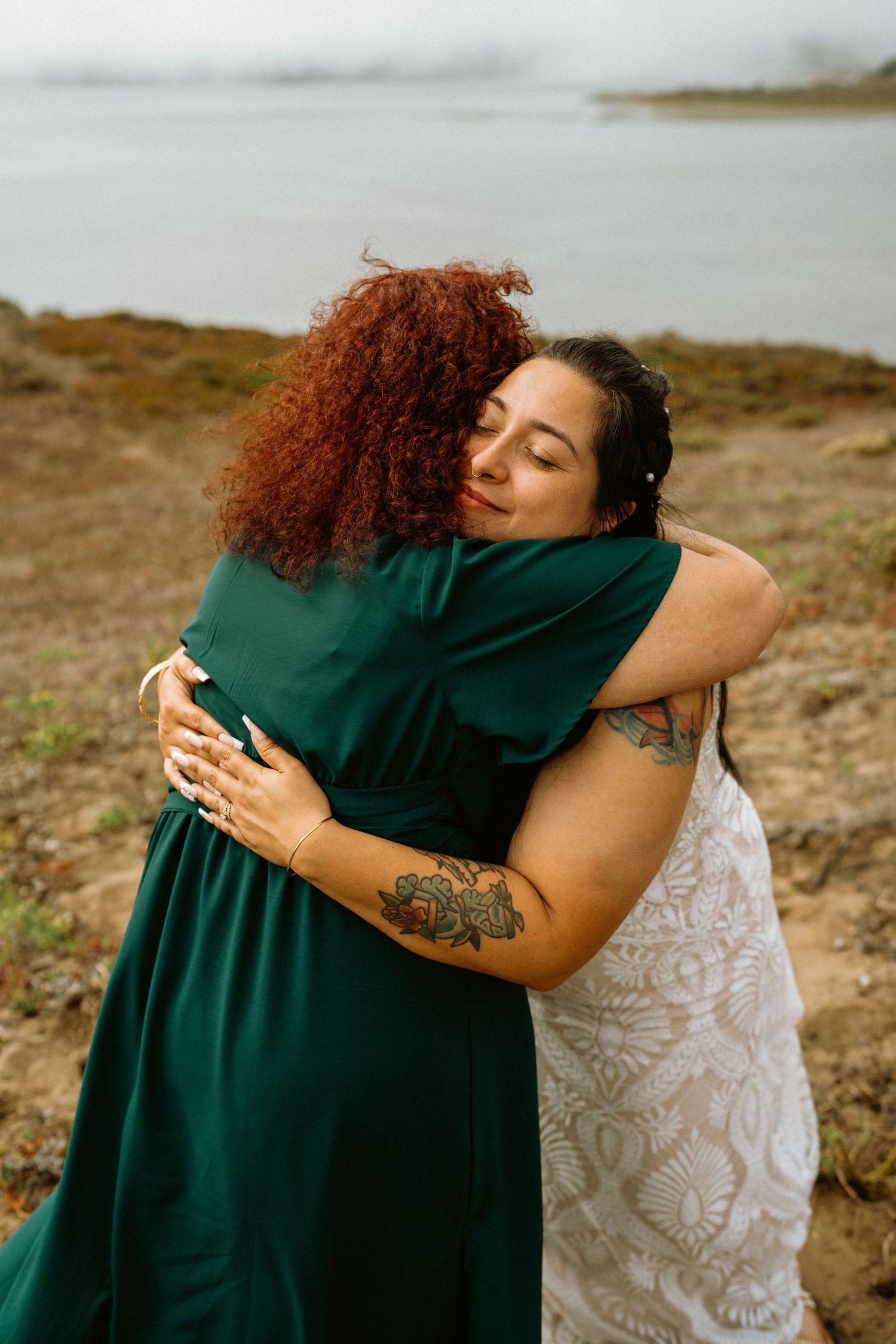 Two women share a warm embrace outdoors near a misty waterfront, one in green dress one in white lace.