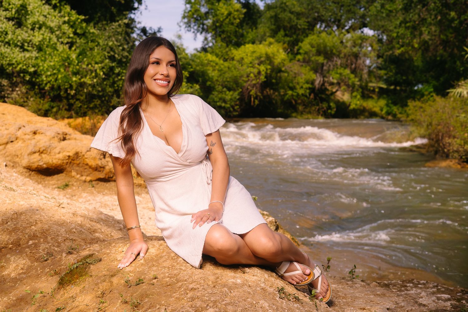 A smiling young woman in a white dress sits on rocks beside a flowing river in a natural setting.