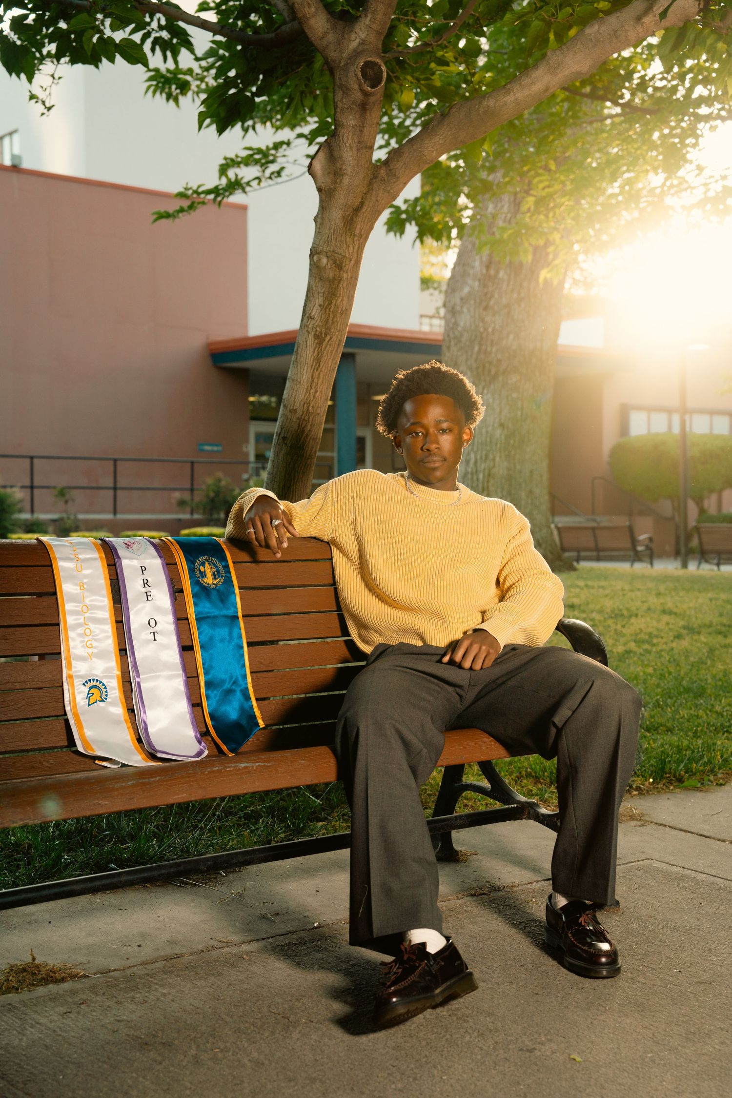 Graduate in yellow sweater sitting on campus bench beside blue and gold graduation stole in golden sunlight.