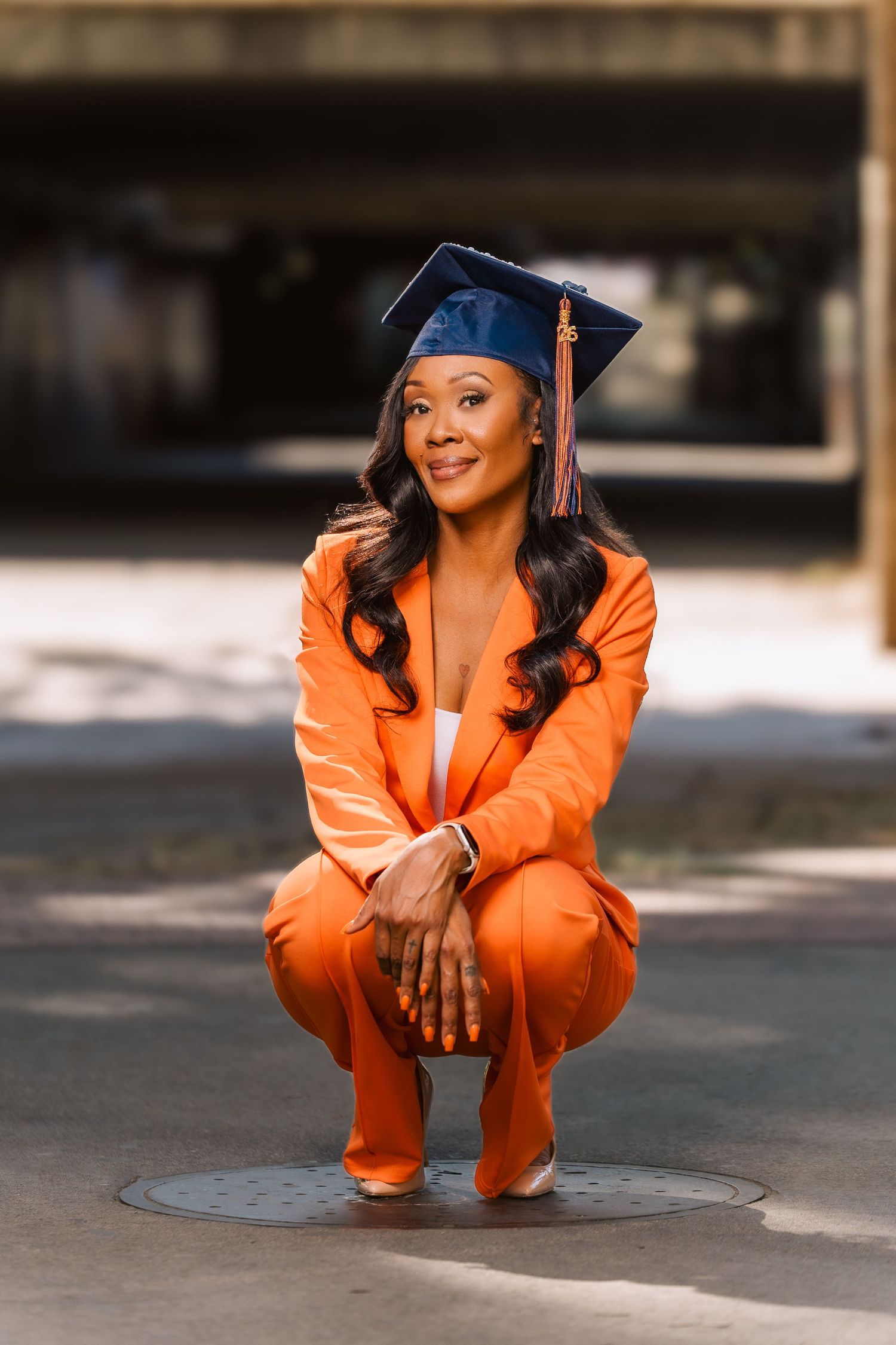 Smiling graduate in orange suit and navy cap crouching low on a pathway in an urban setting.