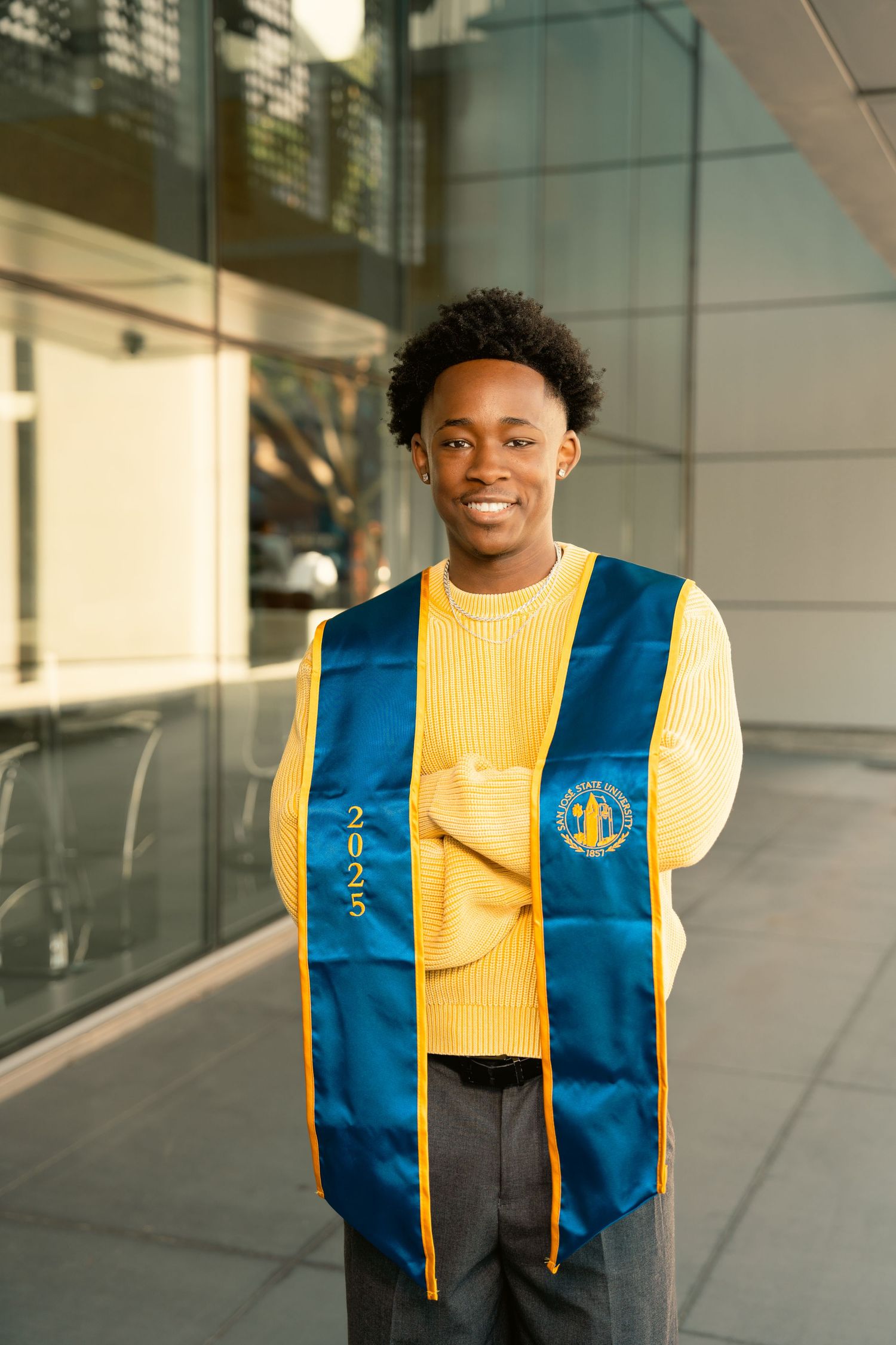 Smiling graduate wearing yellow sweater and blue 2025 graduation stole standing outside glass building.