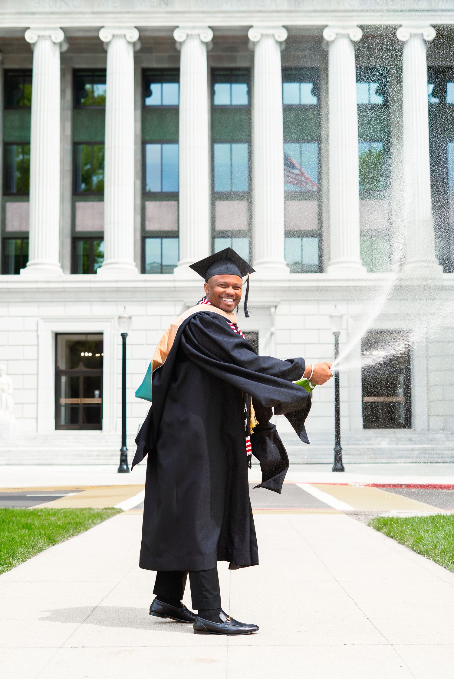 Smiling graduate in cap and gown spraying champagne outside university building.