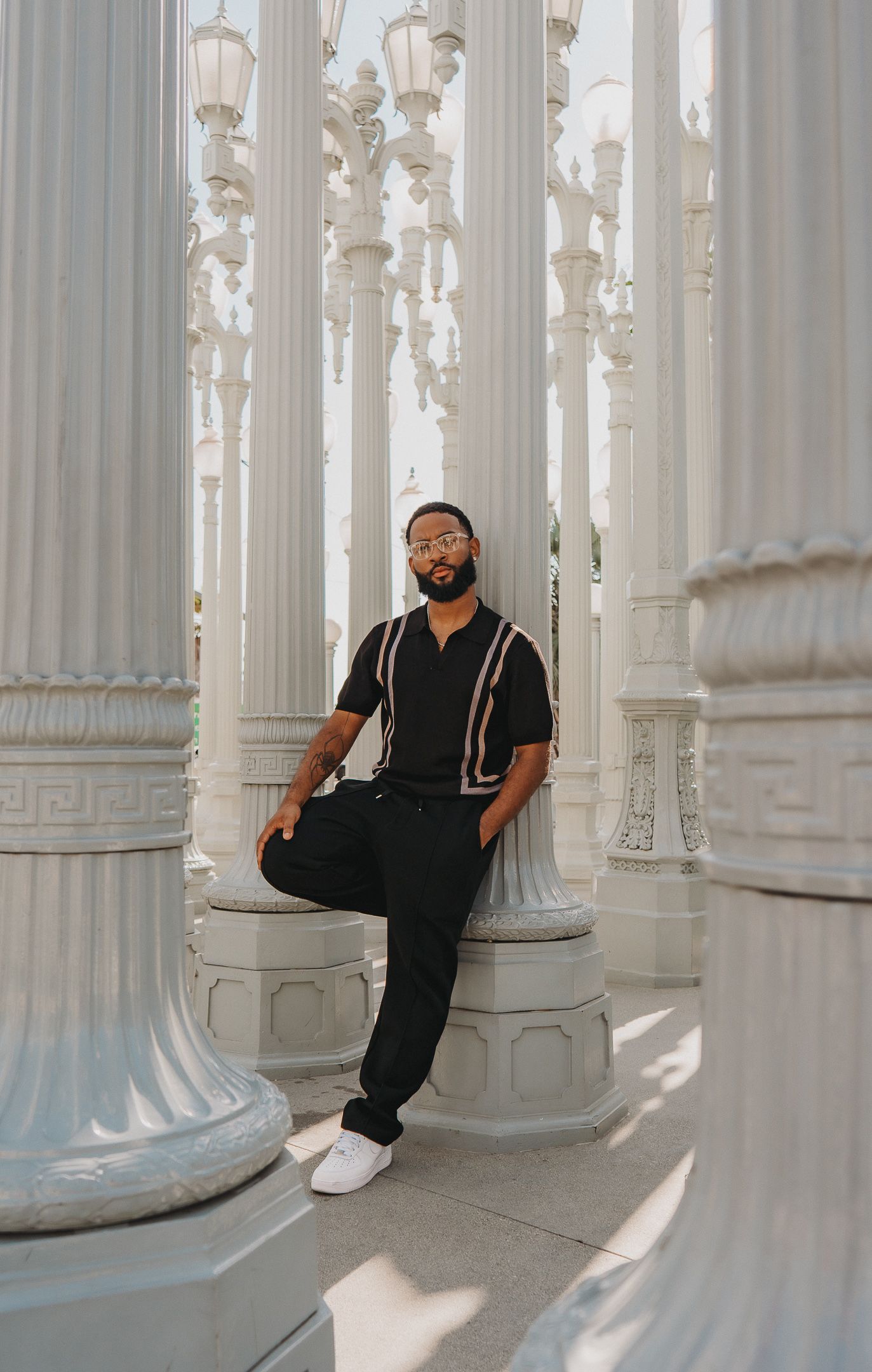 Stylish man in black outfit leaning against white columns at LACMA urban lights.