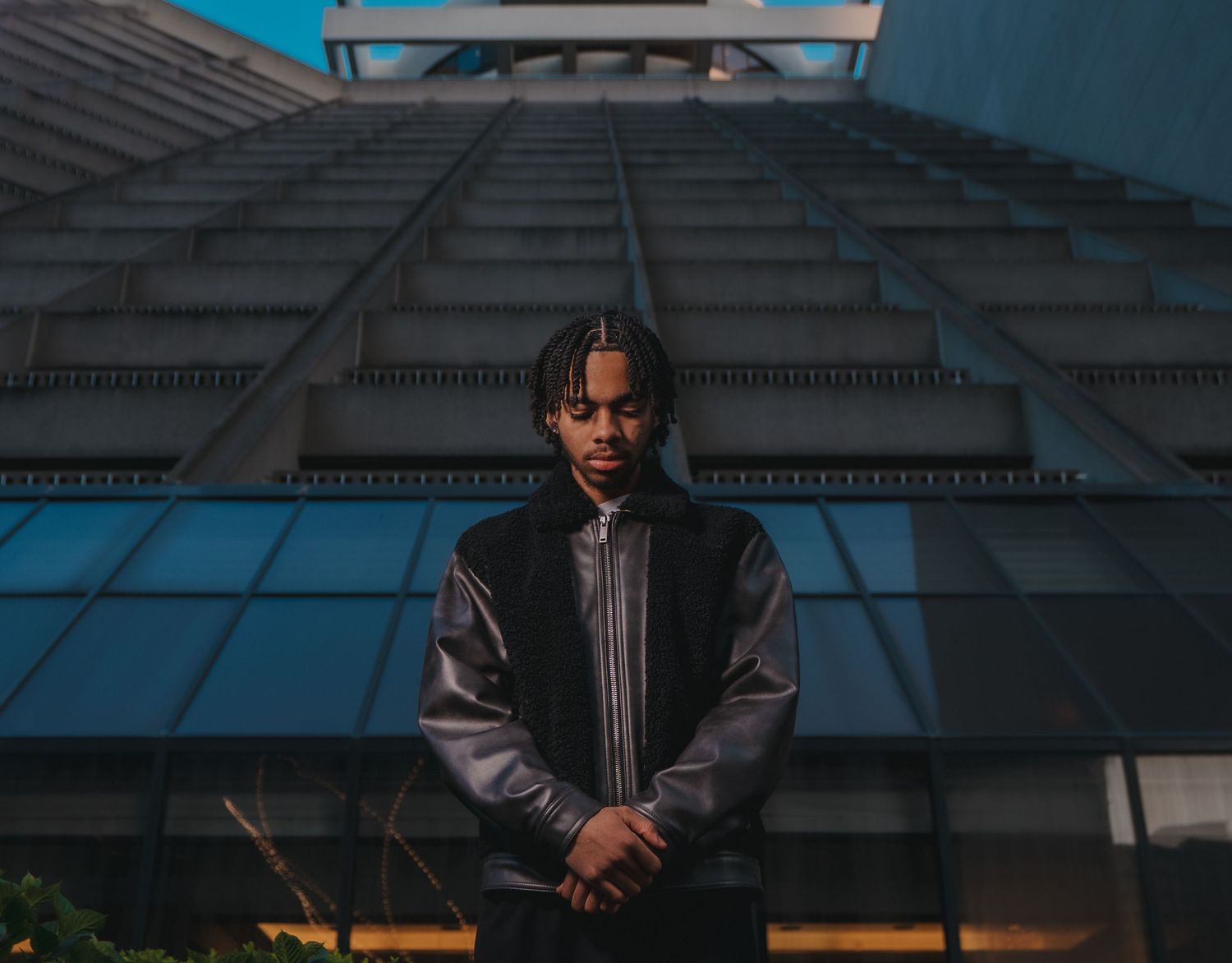 Young man in leather jacket stands confidently before modern glass skyscraper facade.