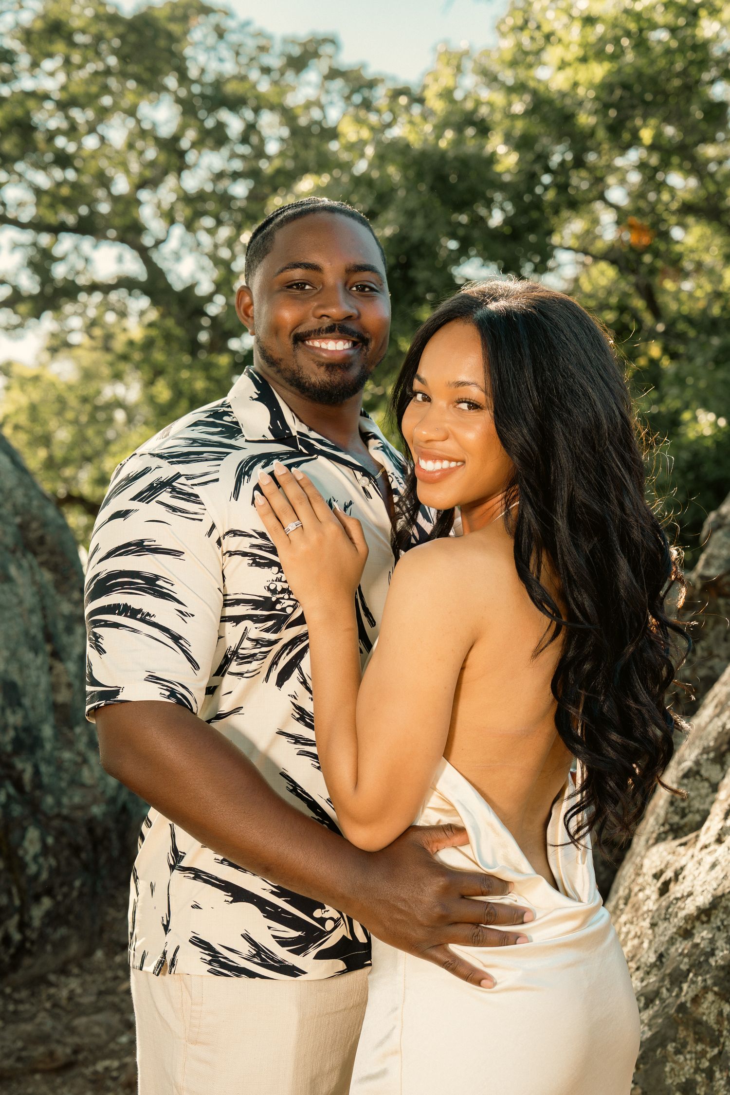 Smiling couple embraces outdoors among rocks and trees during golden hour engagement session.