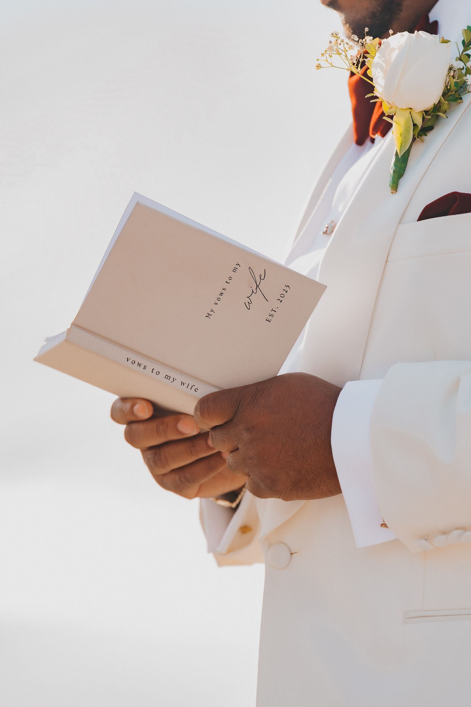 Groom in white tuxedo jacket reading wedding vows from elegant booklet with floral boutonniere.