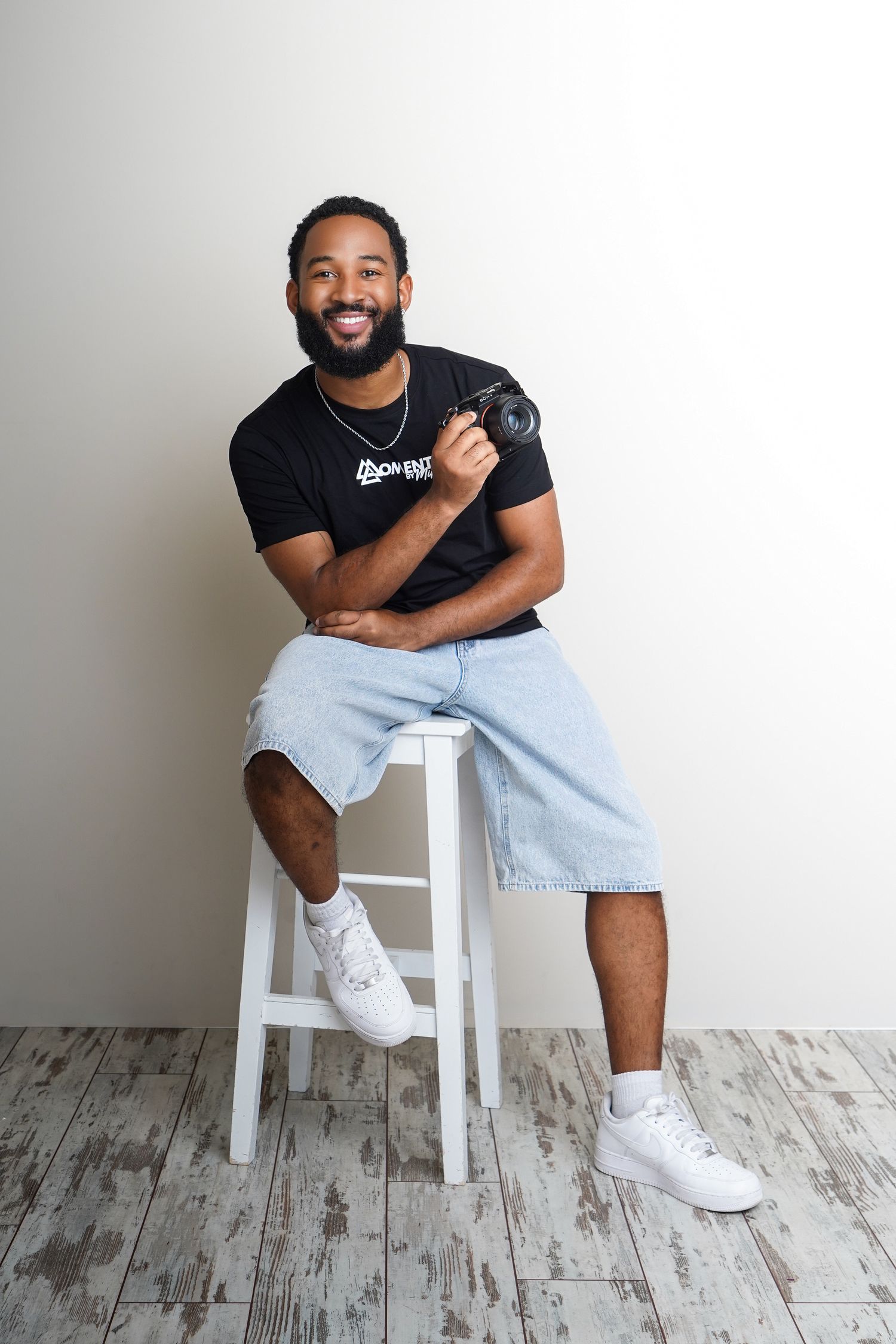 Smiling bearded man holding camera seated on white stool in bright studio setting.
