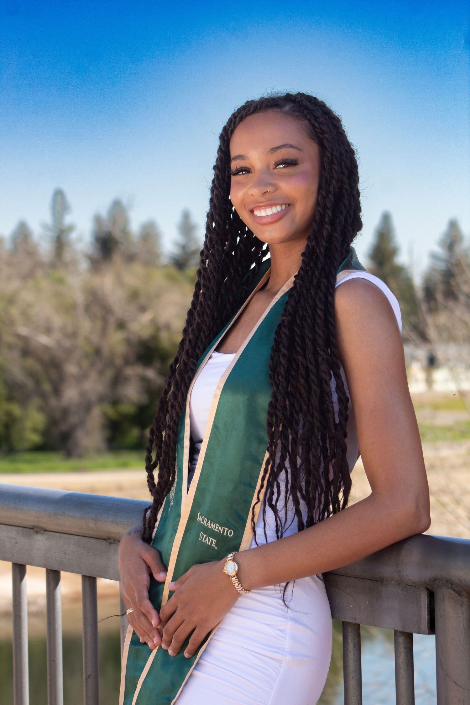 Smiling graduate with long braids wearing white dress and green Sacramento State sash outdoors.