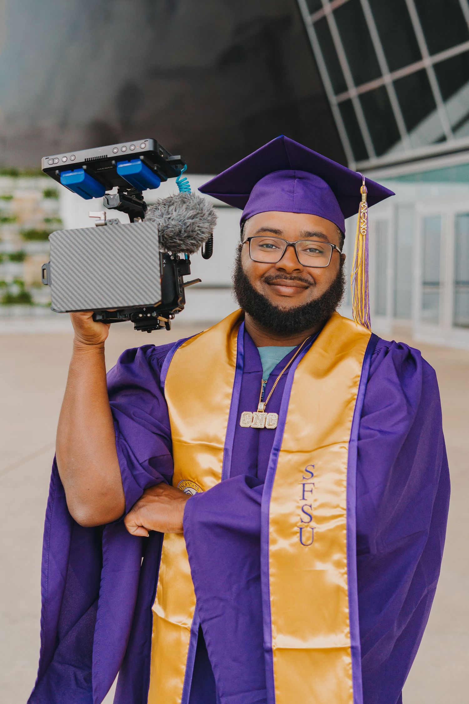 SFSU graduate in purple cap and gold stole holds professional camera rig outside modern building.