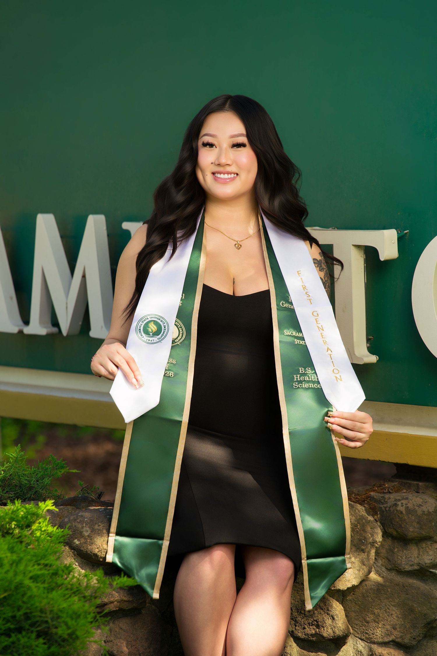 Smiling graduate in black dress displaying green and white graduation stole outdoors.