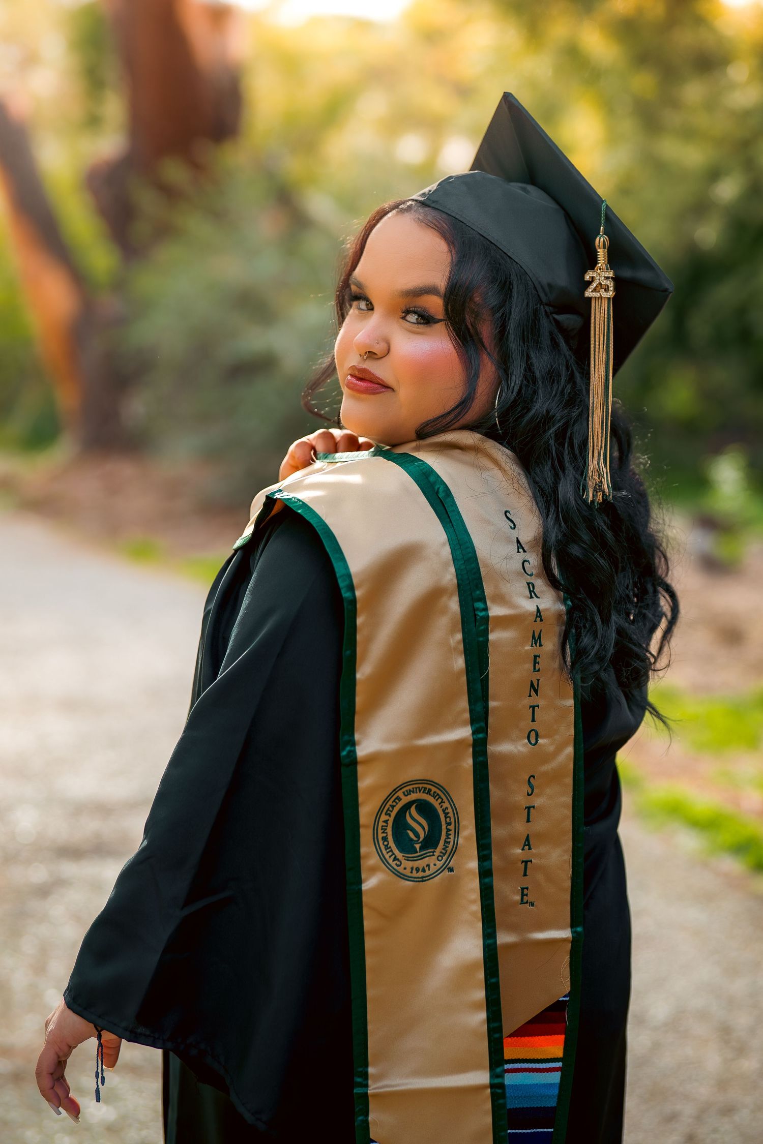 Graduate in black cap and gown with Sacramento State stole posing outdoors at golden hour.
