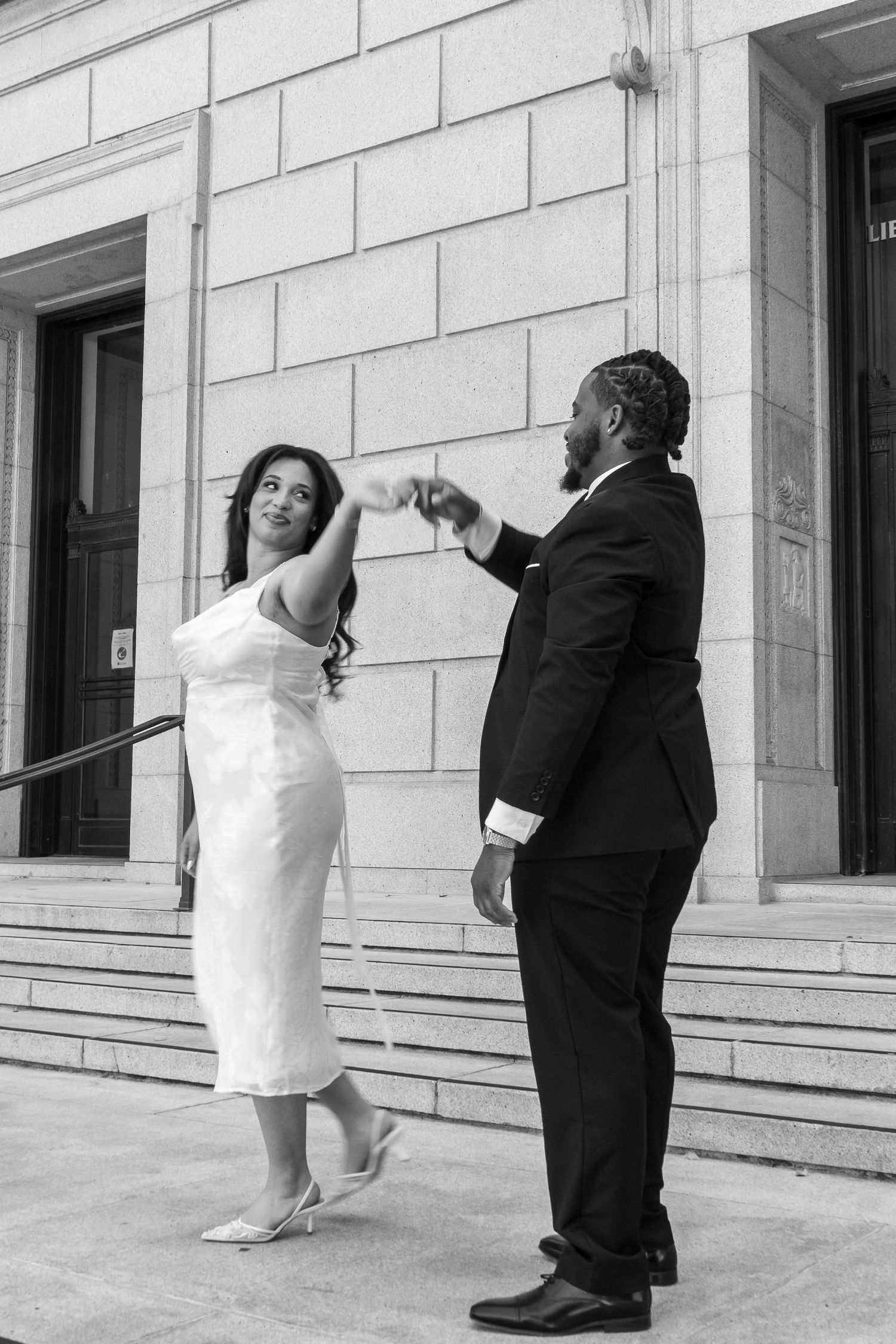 Black and white photo of groom spinning bride in white midi dress on building steps.